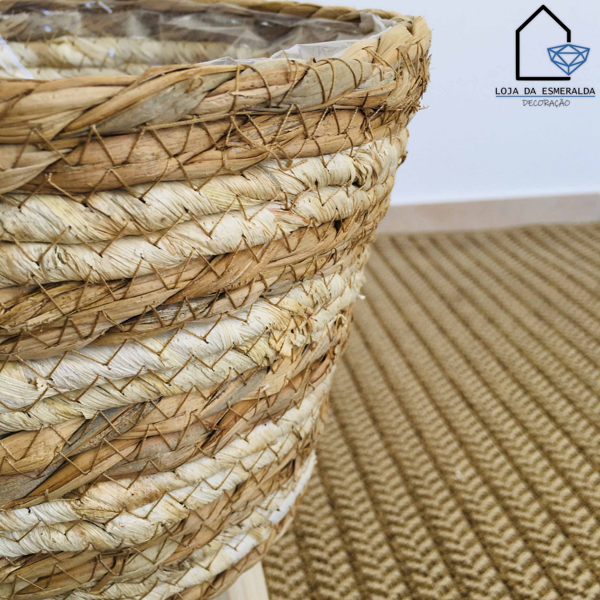Cesto artesanal em fibras naturais com textura trançada em cima de tapete castanho e logotipo LOJA DA ESMERALDA DECORAÇÃO