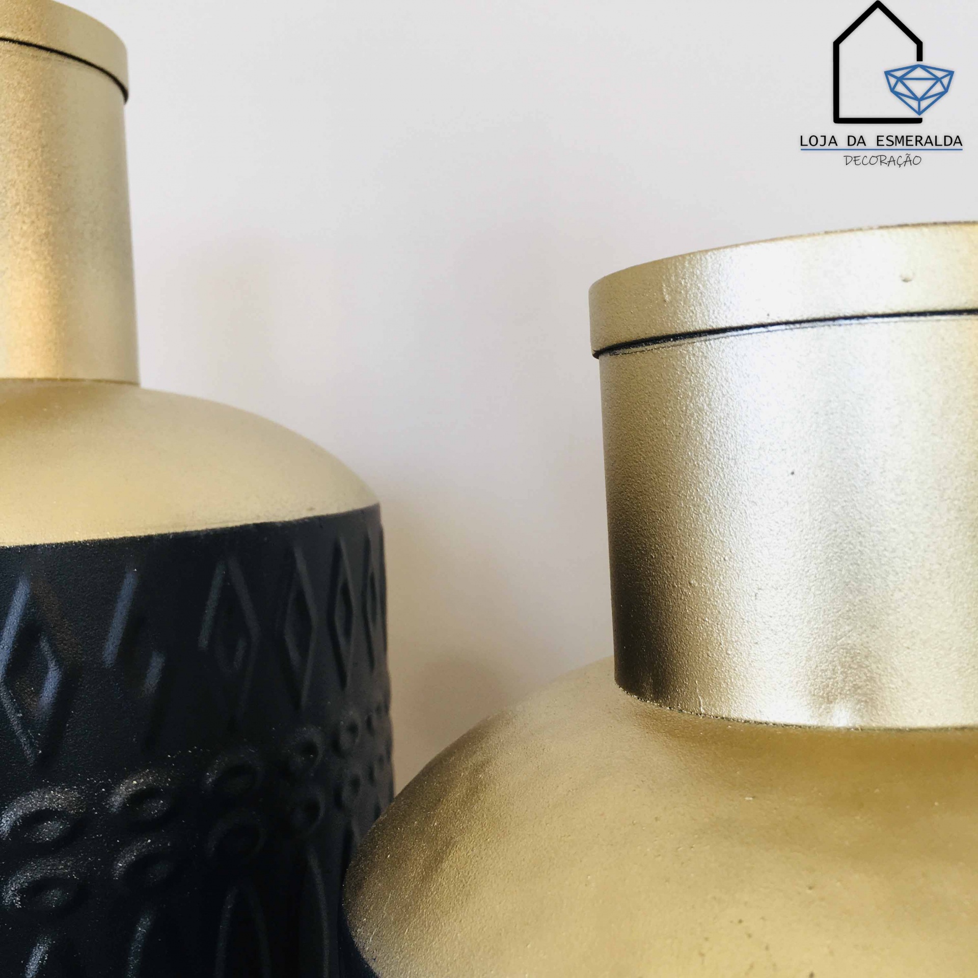Dois vasos decorativos, um preto com padrão geométrico e dourado, outro dourado liso, com logotipo da loja no fundo