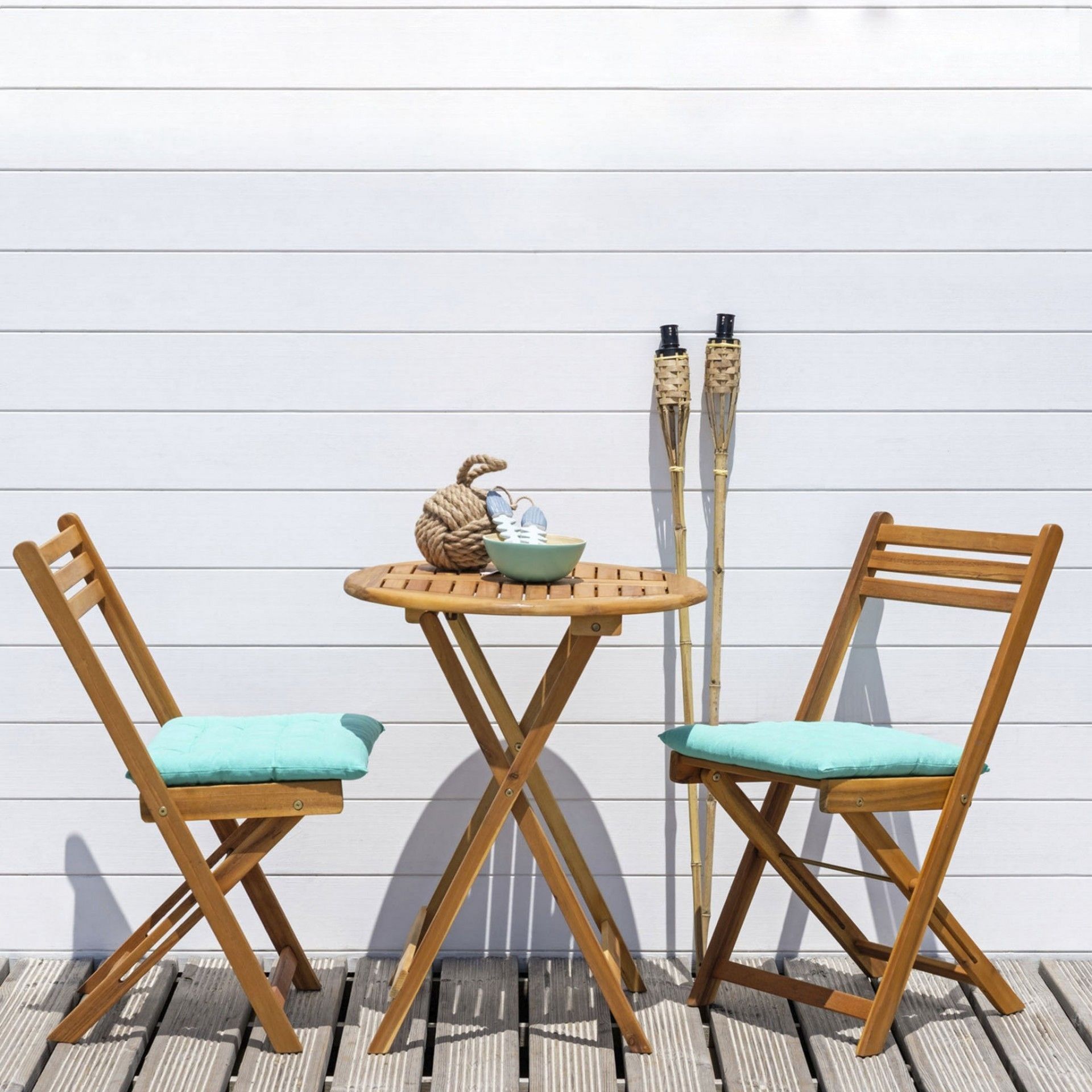 Conjunto de mesa e cadeiras de madeira com almofadas azuis claras num deck em frente a uma parede branca, com tochas de bambu ao lado.