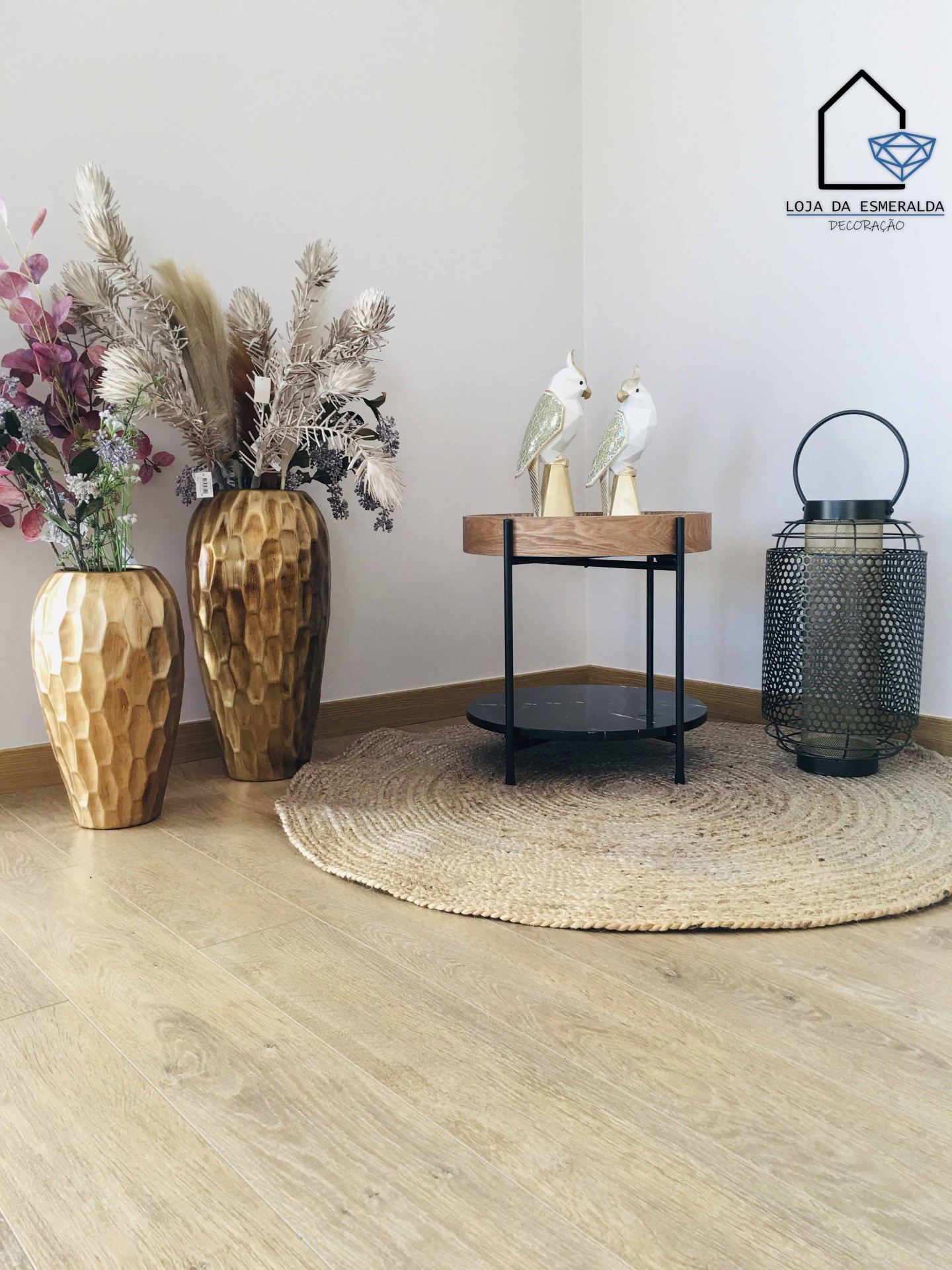 Decoração com vasos dourados, mesa com estatuetas de papagaios, tapete redondo e candeeiro metálico preto num canto com chão em madeira clara