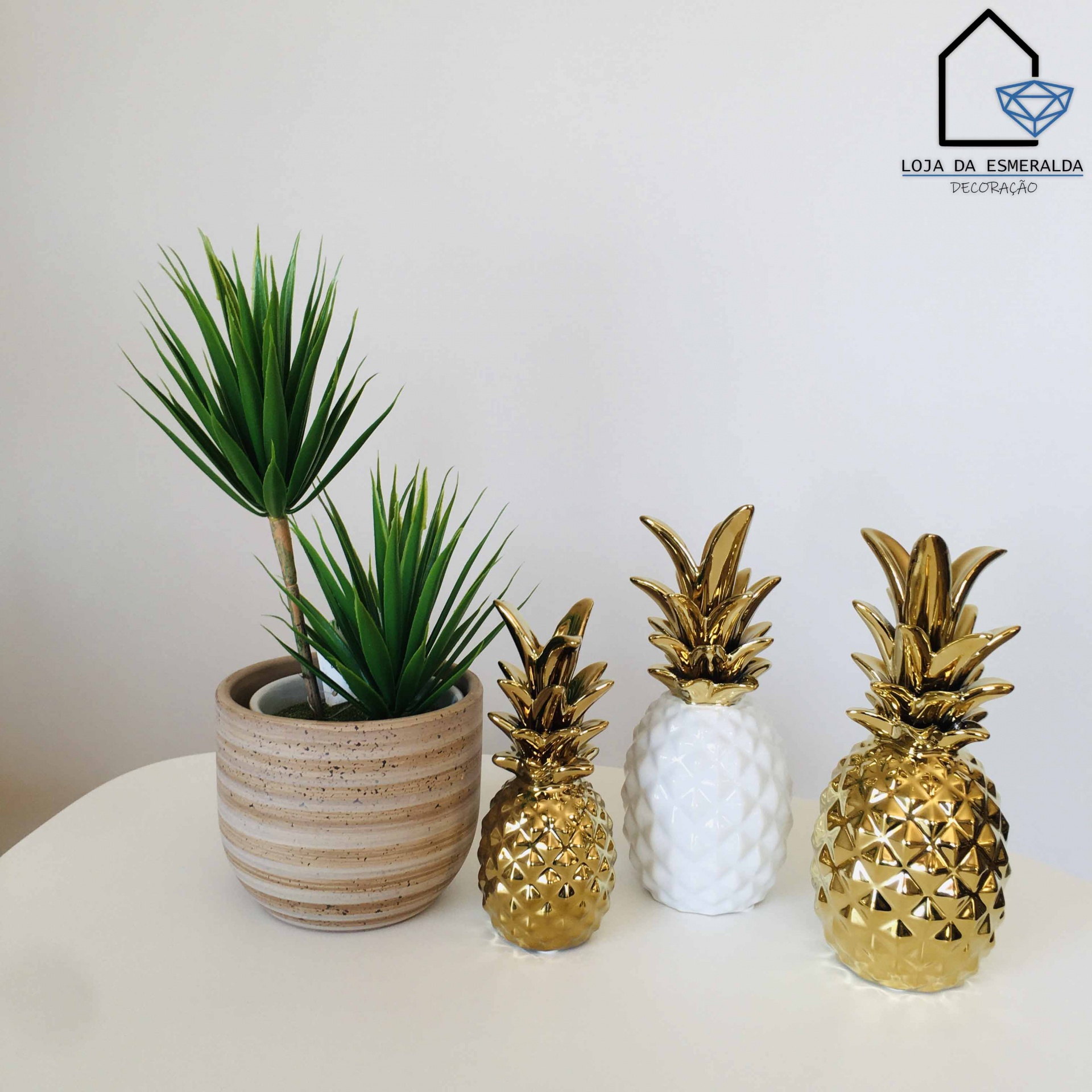 Vaso de cerâmica com planta verde e três ananases decorativos em cerâmica, dois dourados e um branco, sobre mesa branca.