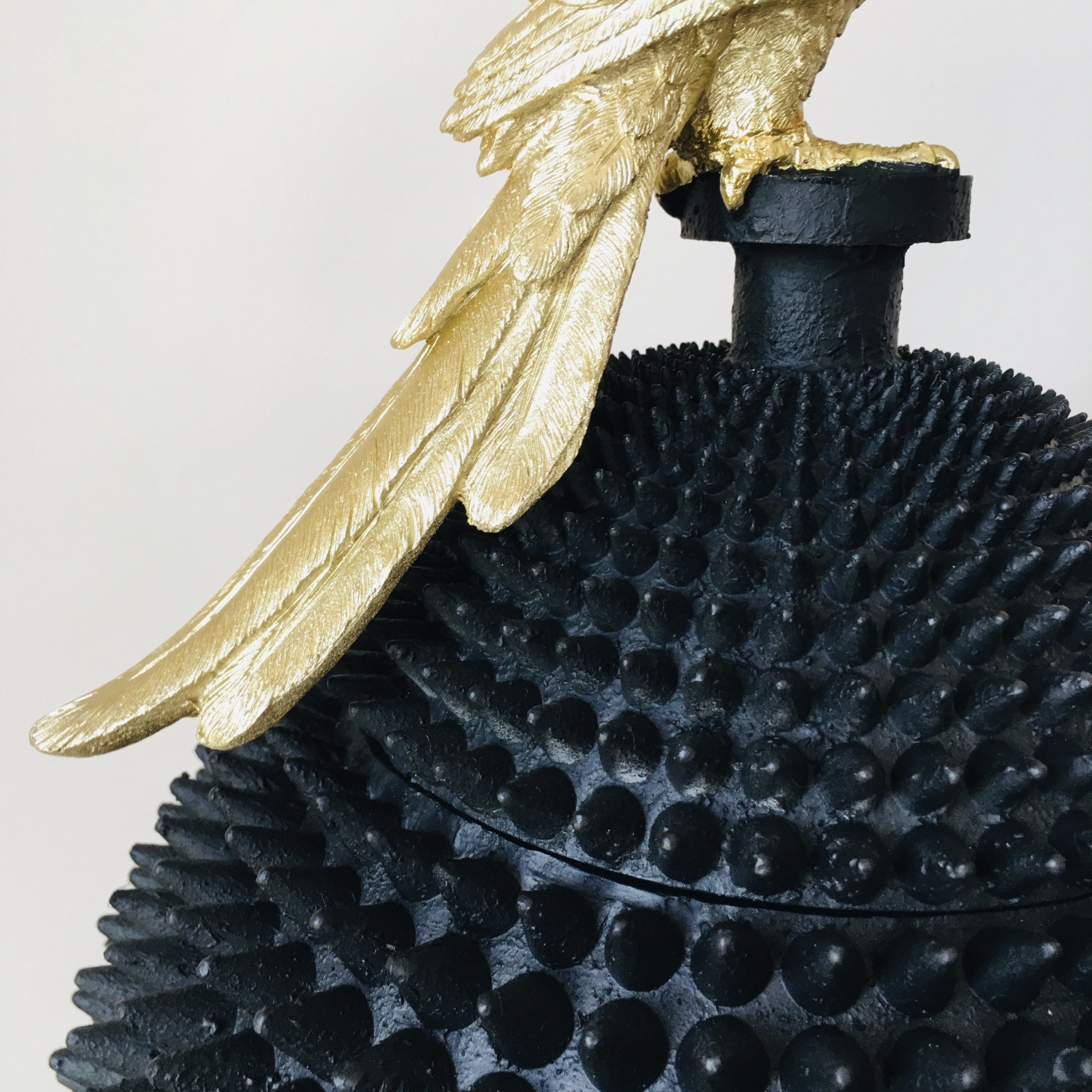 Recipiente decorativo negro com textura espinhosa e tampa com asa dourada de pássaro