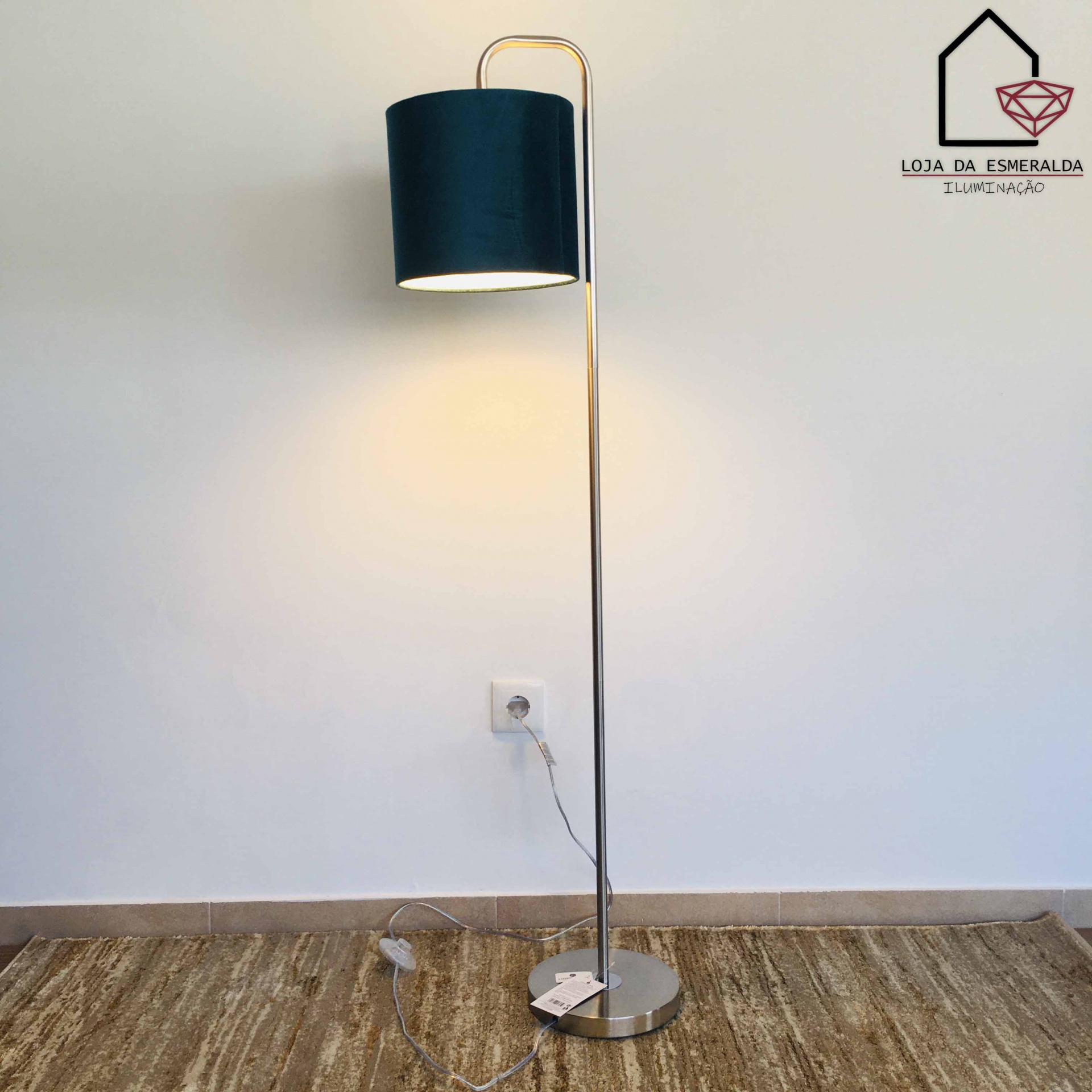 Candeeiro de pé metálico com cúpula azul escura sobre tapete castanho claro junto a parede branca com logo da loja no canto