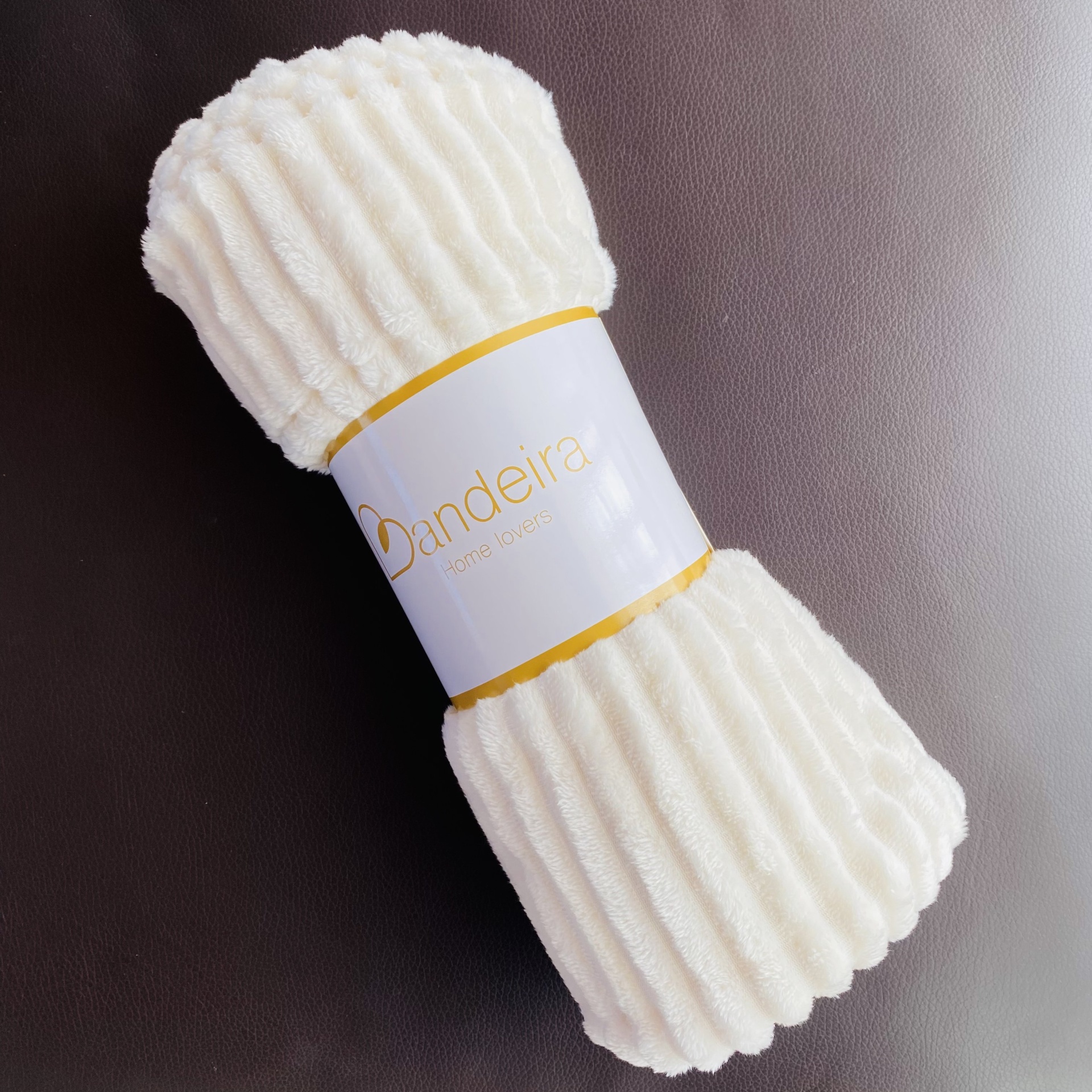 Manta branca felpuda com etiqueta Dandeira Textil Home sobre superfície escura