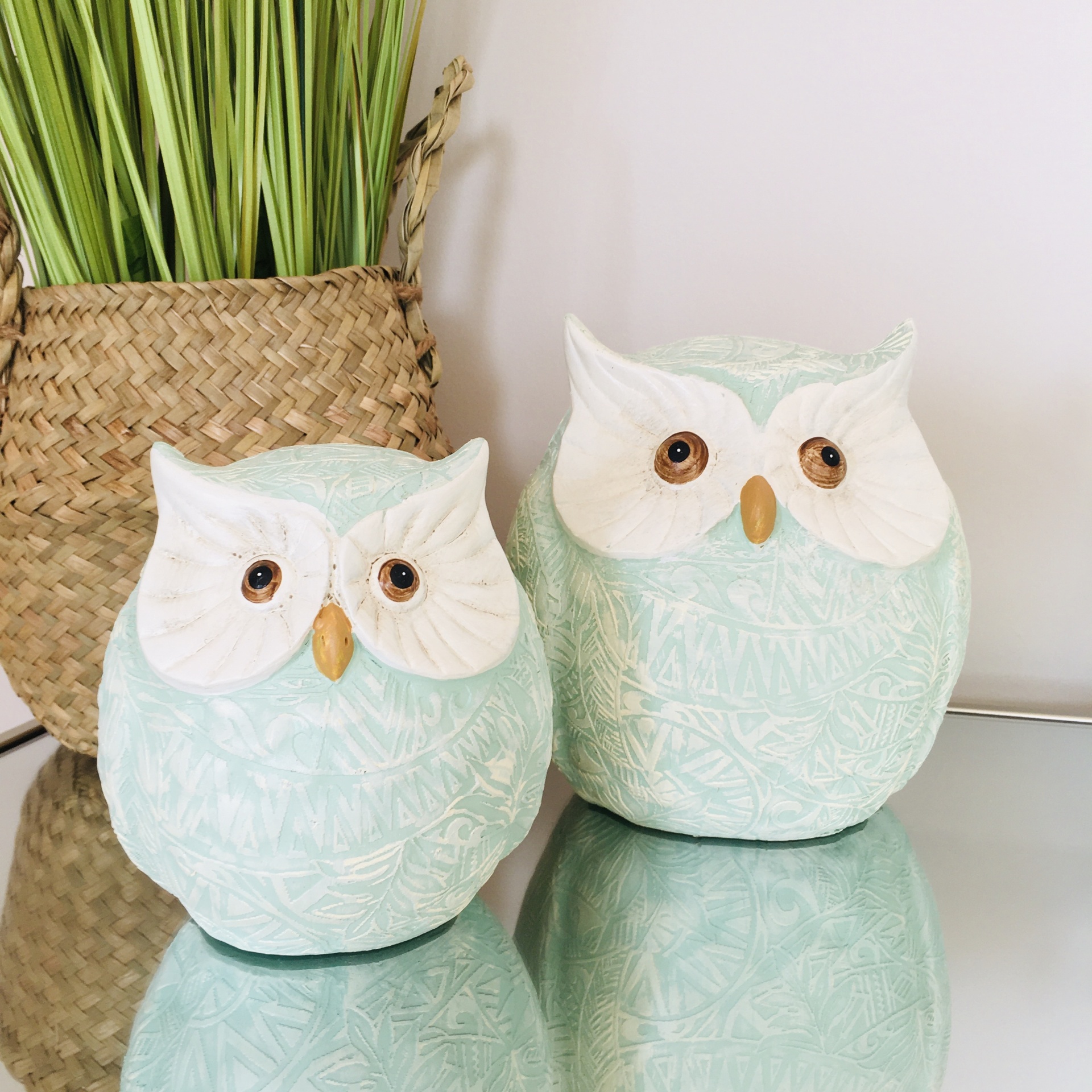 Duas corujas decorativas azul claro e branco com olhos castanhos sobre superfície espelhada ao lado de vaso com planta