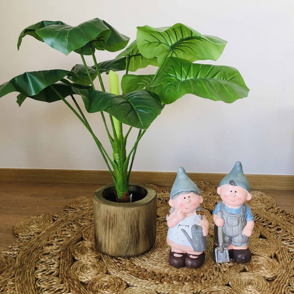 Planta artificial em vaso castanho com dois bonecos de jardim cerâmicos em roupa azul sobre uma toalha redonda de palha.