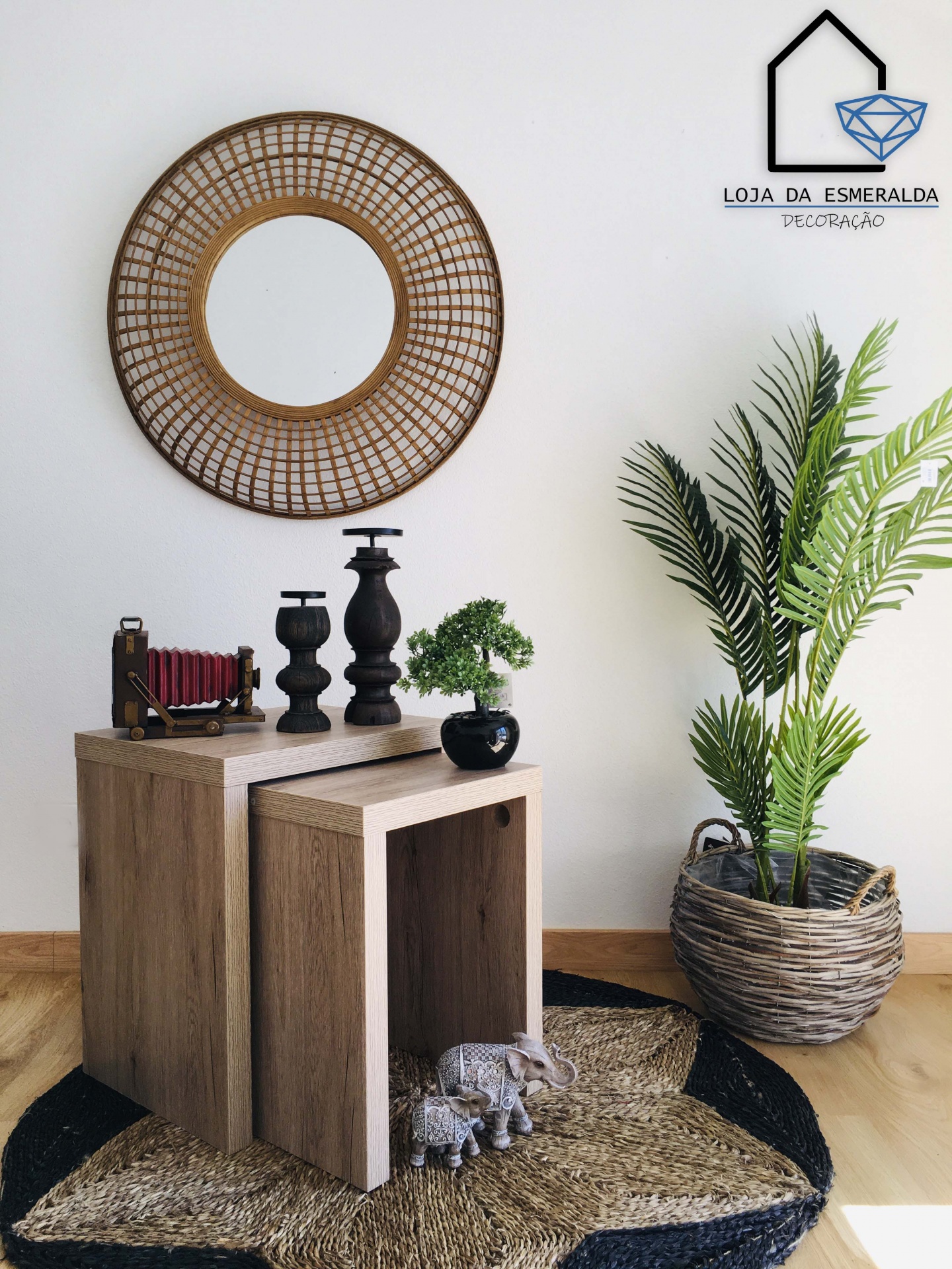 Espelho redondo de rattan, mesas ninho de madeira clara com decoração, plantas artificiais, tapete e logotipo em parede branca.