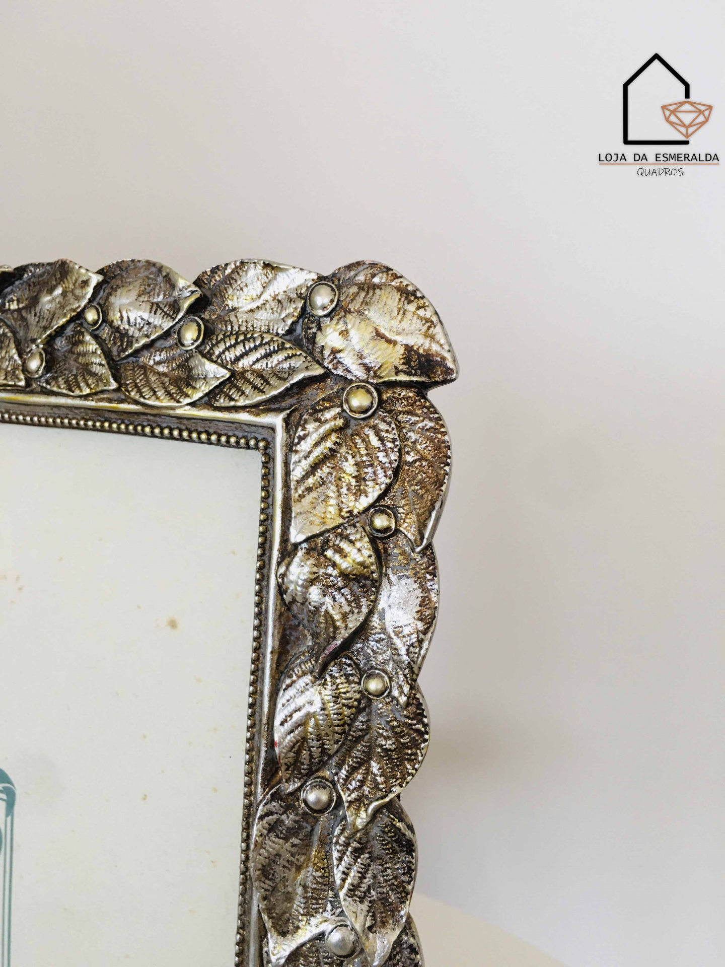 Moldura prateada com folhas texturizadas e detalhes redondos, sobre fundo branco com logotipo Loja da Esmeralda Quartos