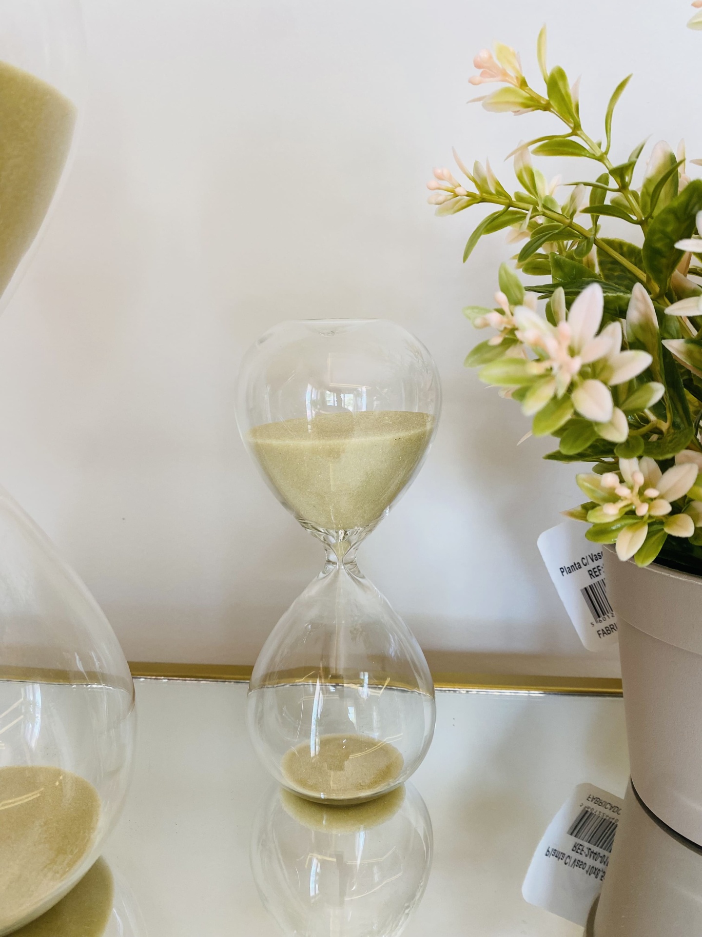 Ampulheta de vidro com areia bege ao lado de vaso branco com planta artificial