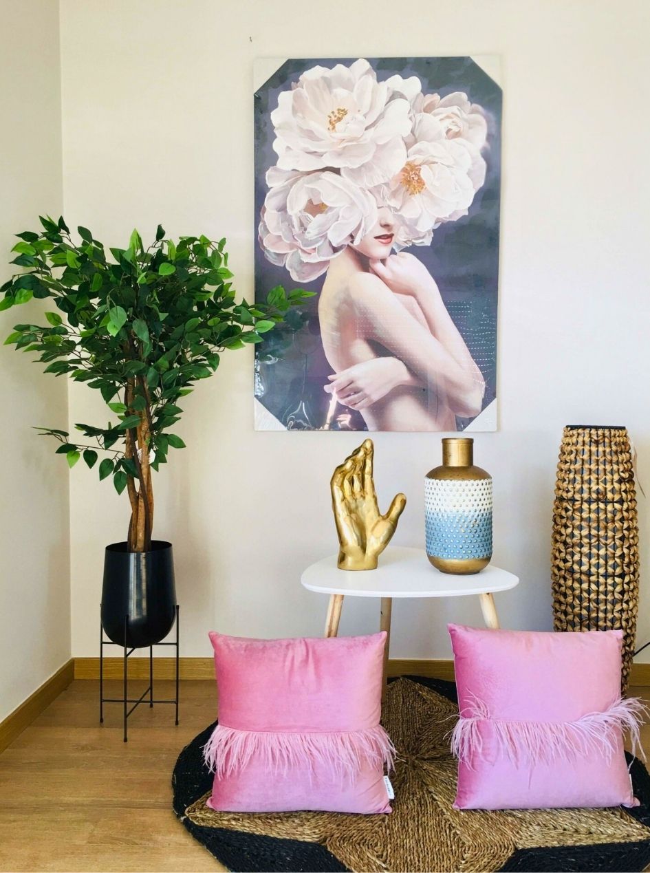 Decoração de sala com almofadas rosas, mesa com decoração dourada e azul, planta em vaso preto, tapete natural e quadro artístico de mulher