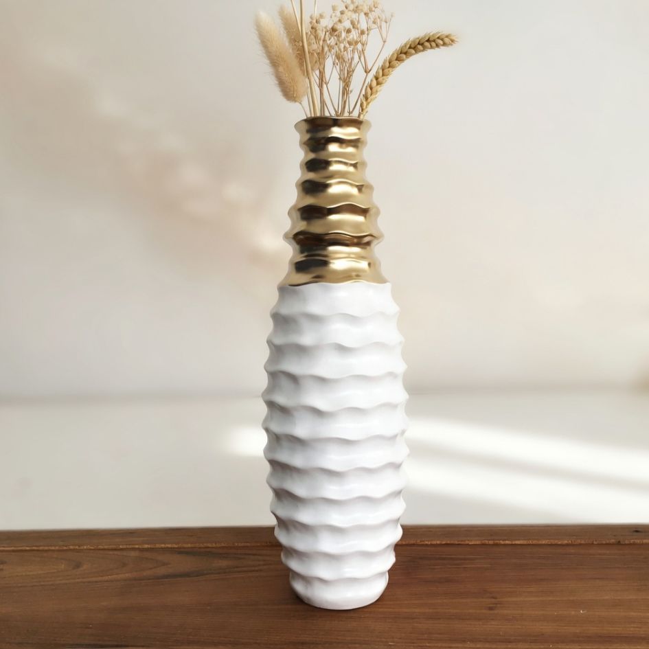 Vaso de cerâmica branco e dourado com textura ondulada, numa mesa de madeira com ramos secos.