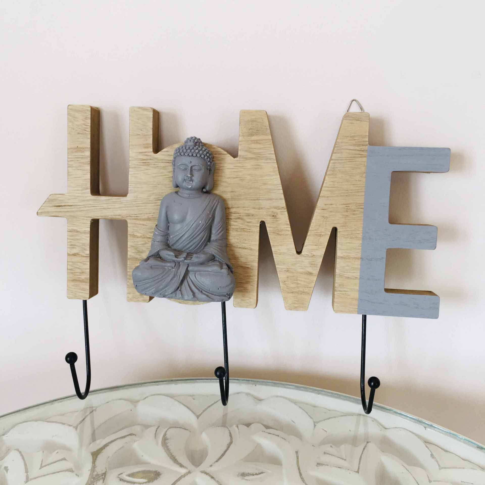 Cabide de parede com a palavra HOME em madeira e Buddha cinzento integrado na letra O e três ganchos de metal preto.