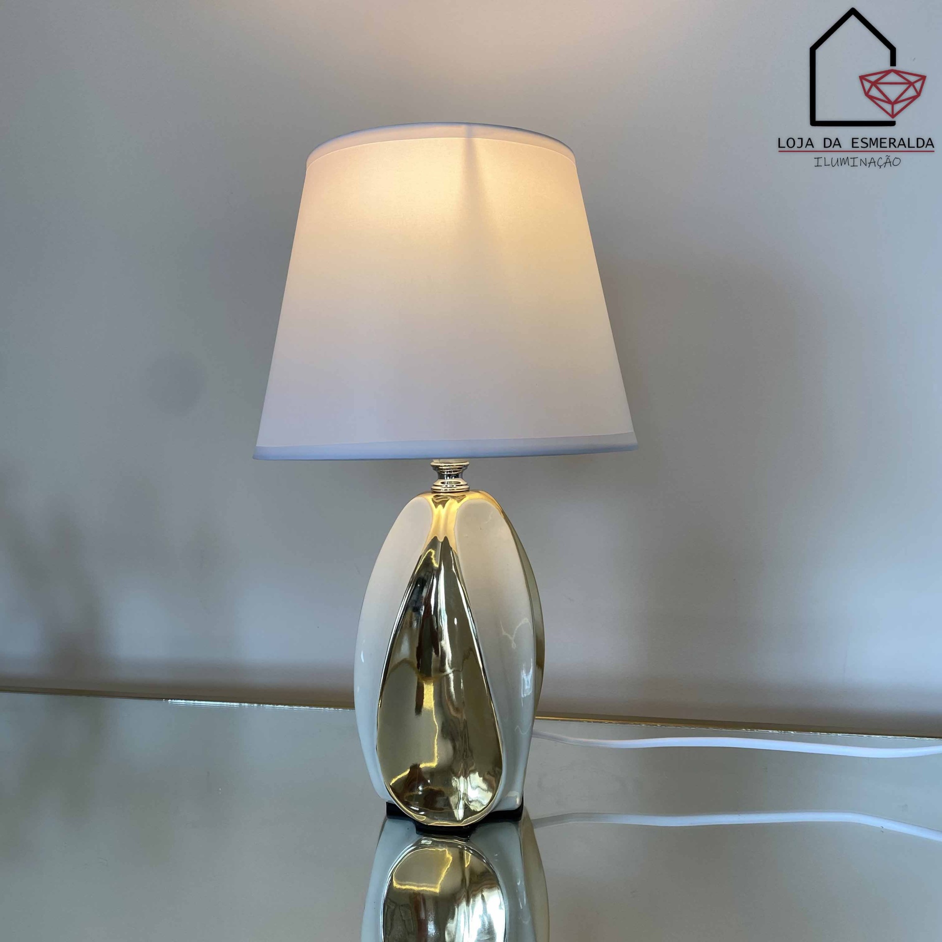 Candeeiro de mesa com base dourada e sombra branca numa superfície espelhada