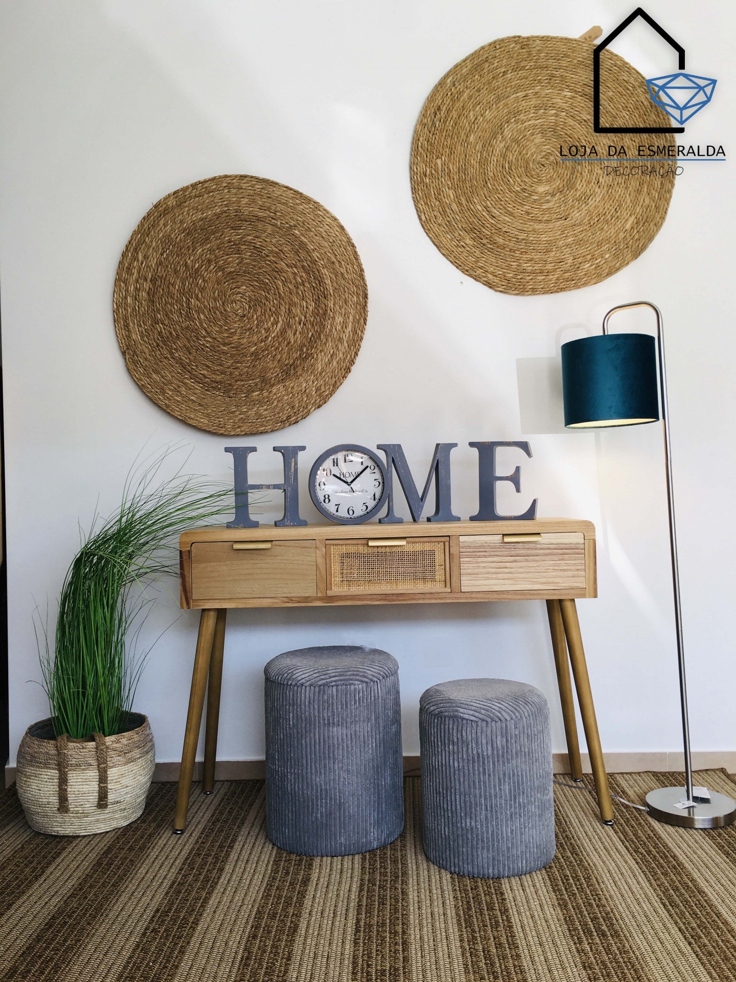 Conjunto de mobília com mesa de madeira, dois pufes cinzentos, candeeiro de pé azul, vaso com planta e cestarias redondas na parede