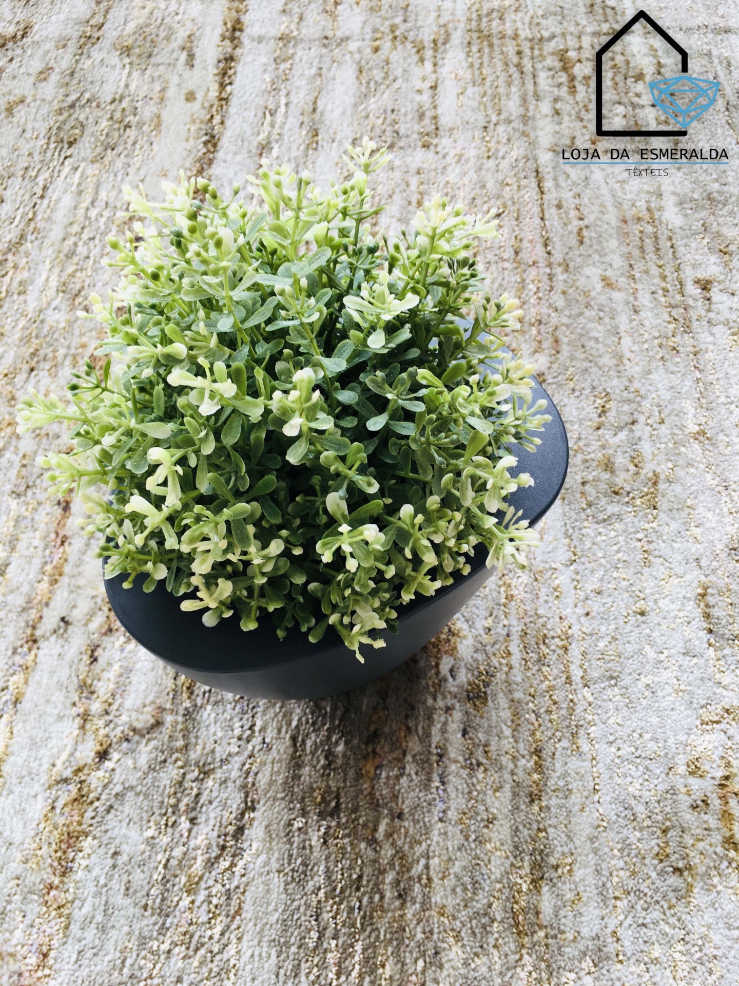Planta decorativa artificial em vaso preto sobre superfície bege, com texto Loja da Esmeralda Textiles.