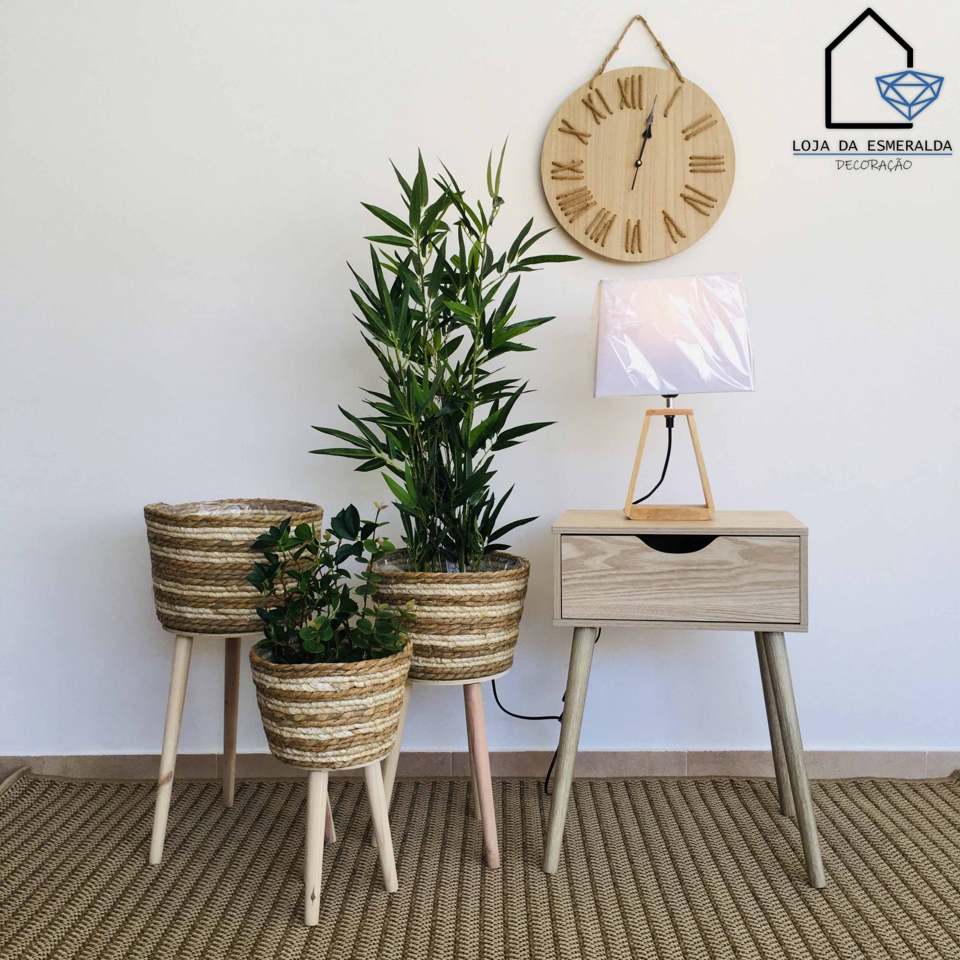 Relógio de parede, vasos de plantas em suportes de ráfia e madeira, candeeiro e mesa de cabeceira em ambiente minimalista