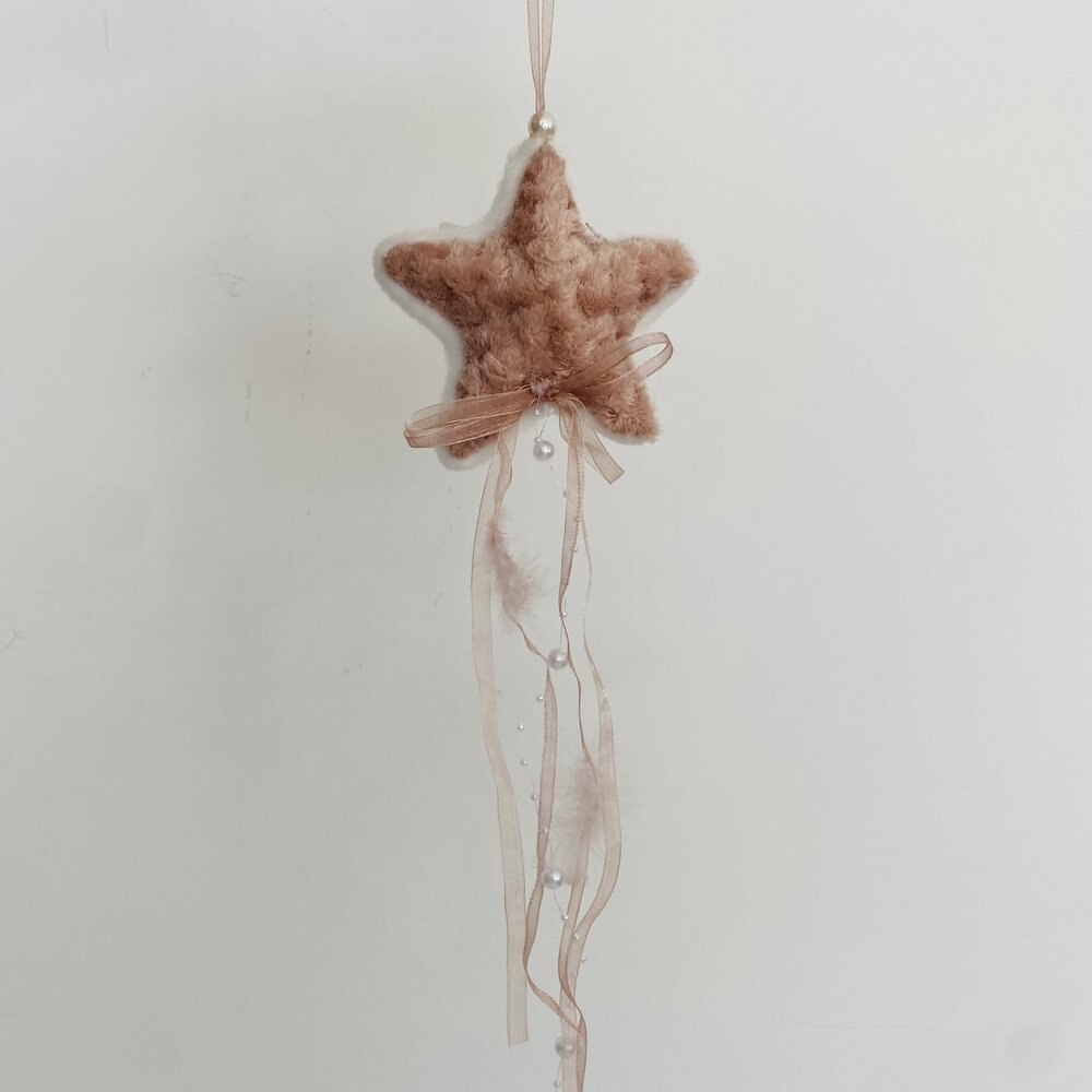 Estrela decorativa de tecido macio castanho claro com fitas e pompons brancos pendurados.