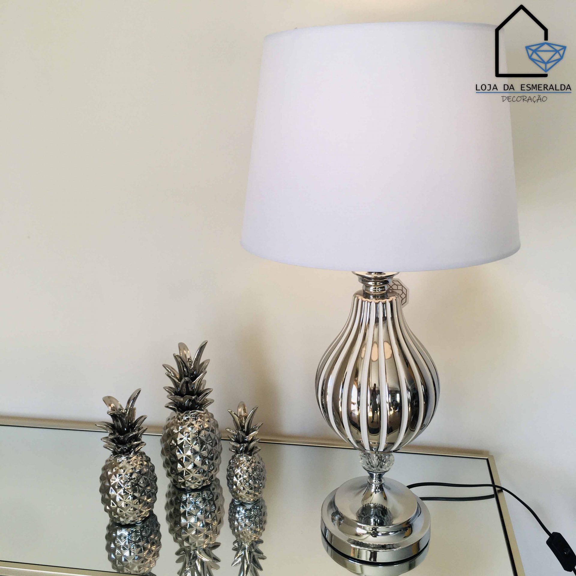 Abajur prateado com cúpula branca e três abacaxis decorativos prateados numa superfície espelhada