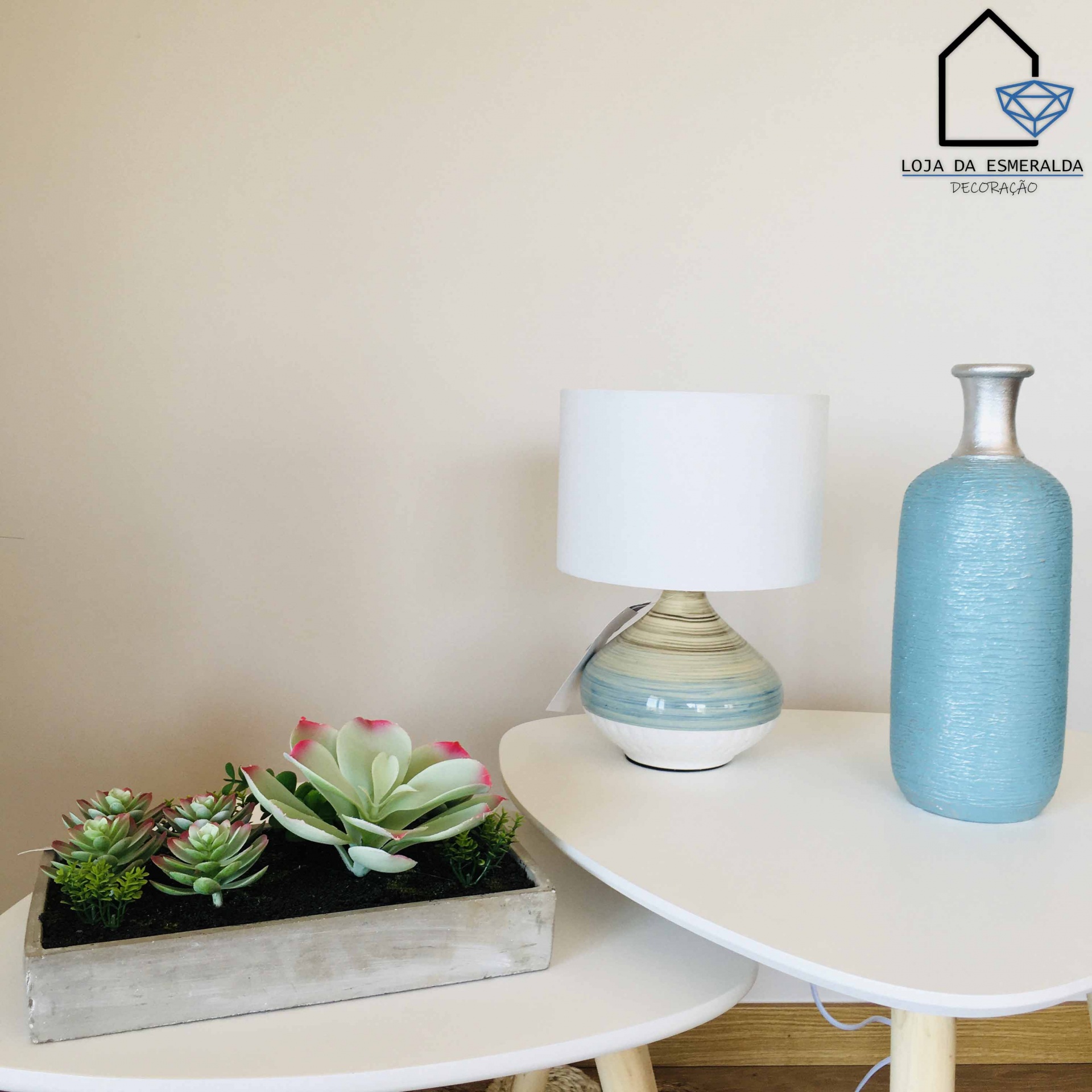 Duas mesas brancas com vaso azul, candeeiro e plantas suculentas, parede bege e logo de loja