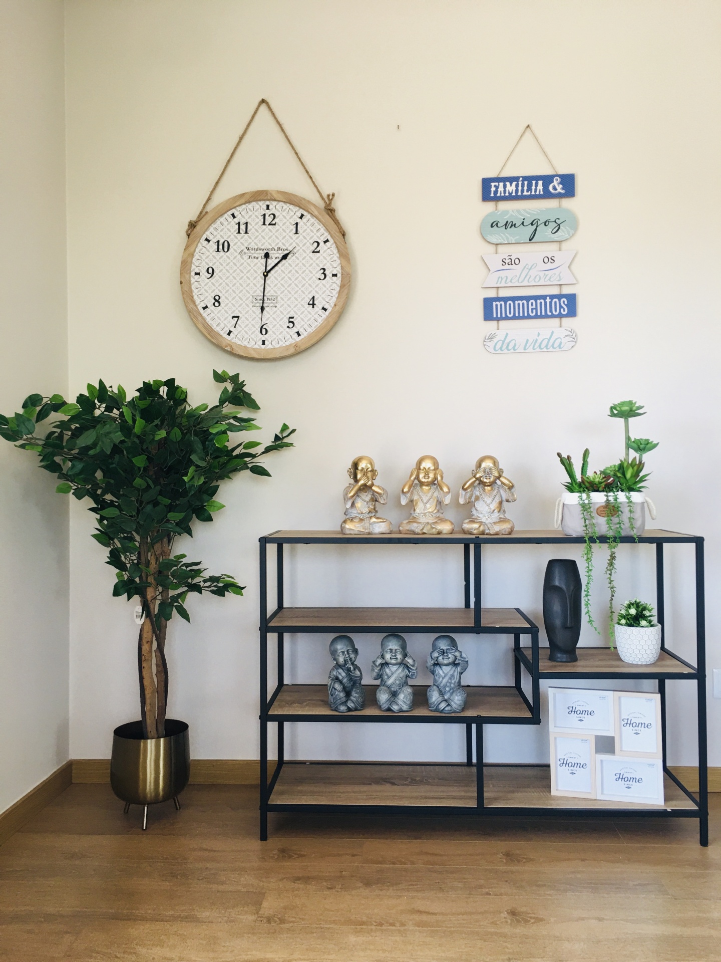 Relógio de parede redondo com moldura em corda, estante com estatuetas e plantas em ambiente com chão de madeira