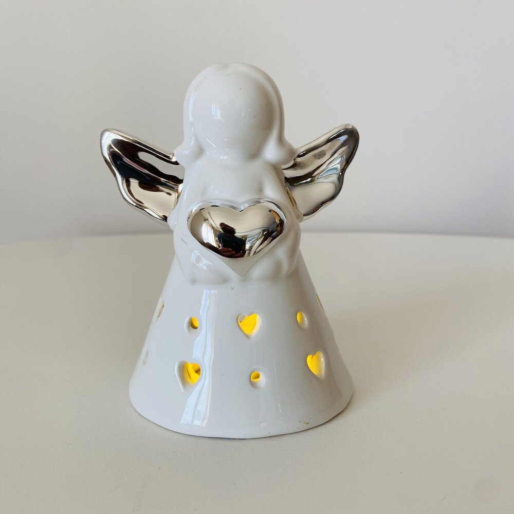Figura decorativa de anjo branco com asas prateadas e luz amarela a passar por recortes em forma de coração