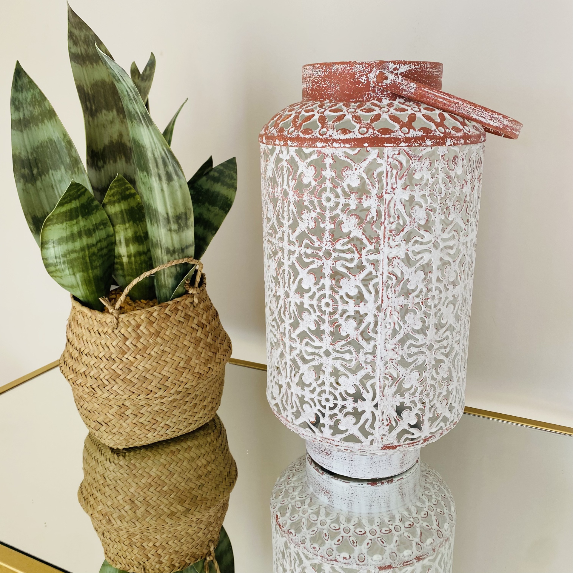 Lanterna metálica decorativa envelhecida com padrão calado, ao lado de planta em vaso de palha