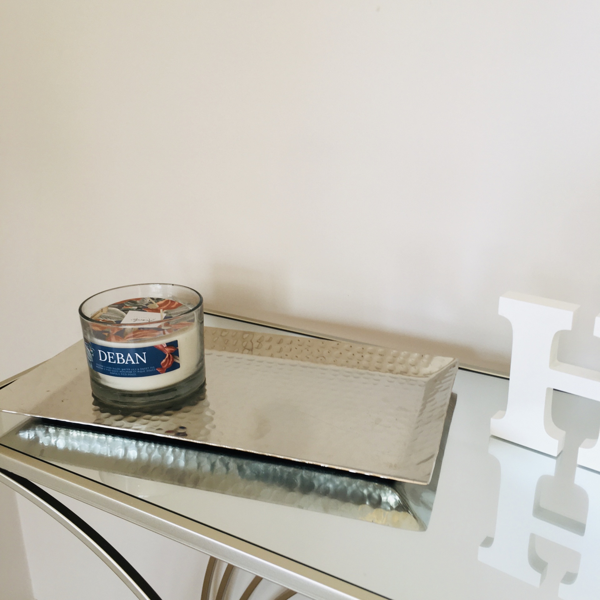 Vela DEBAN em vidro sobre bandeja prateada martelada numa mesa espelhada com letra H branca ao lado