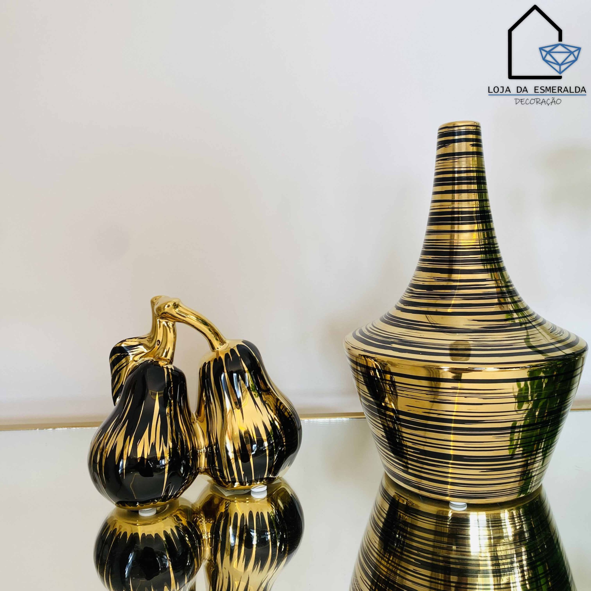 Vaso e dupla peça decorativa dourados com linhas pretas sobre superfície espelhada e fundo branco com logo