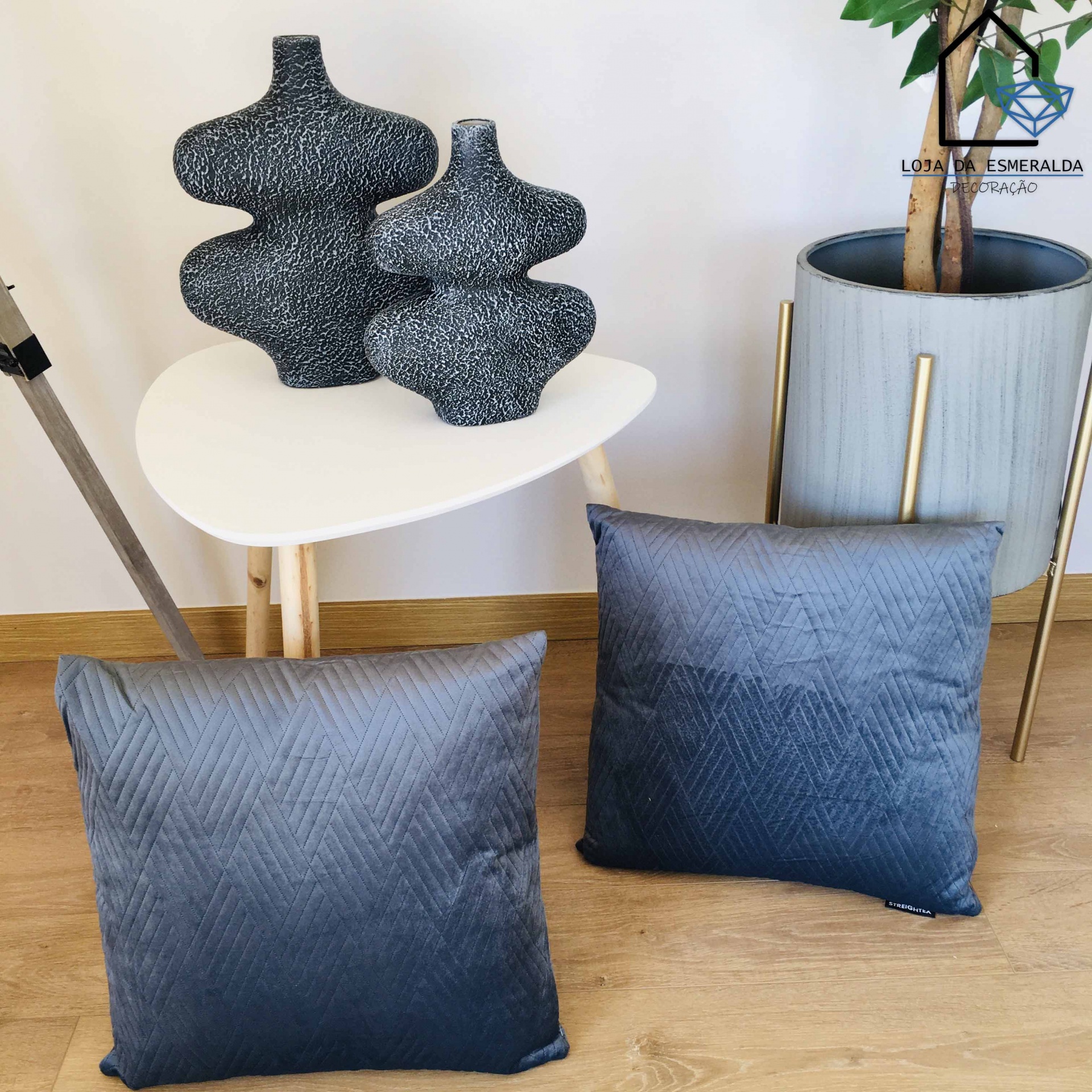 Almofadas azuis com padrão geométrico junto a esculturas decorativas pretas sobre mesa branca e vaso cinza com planta