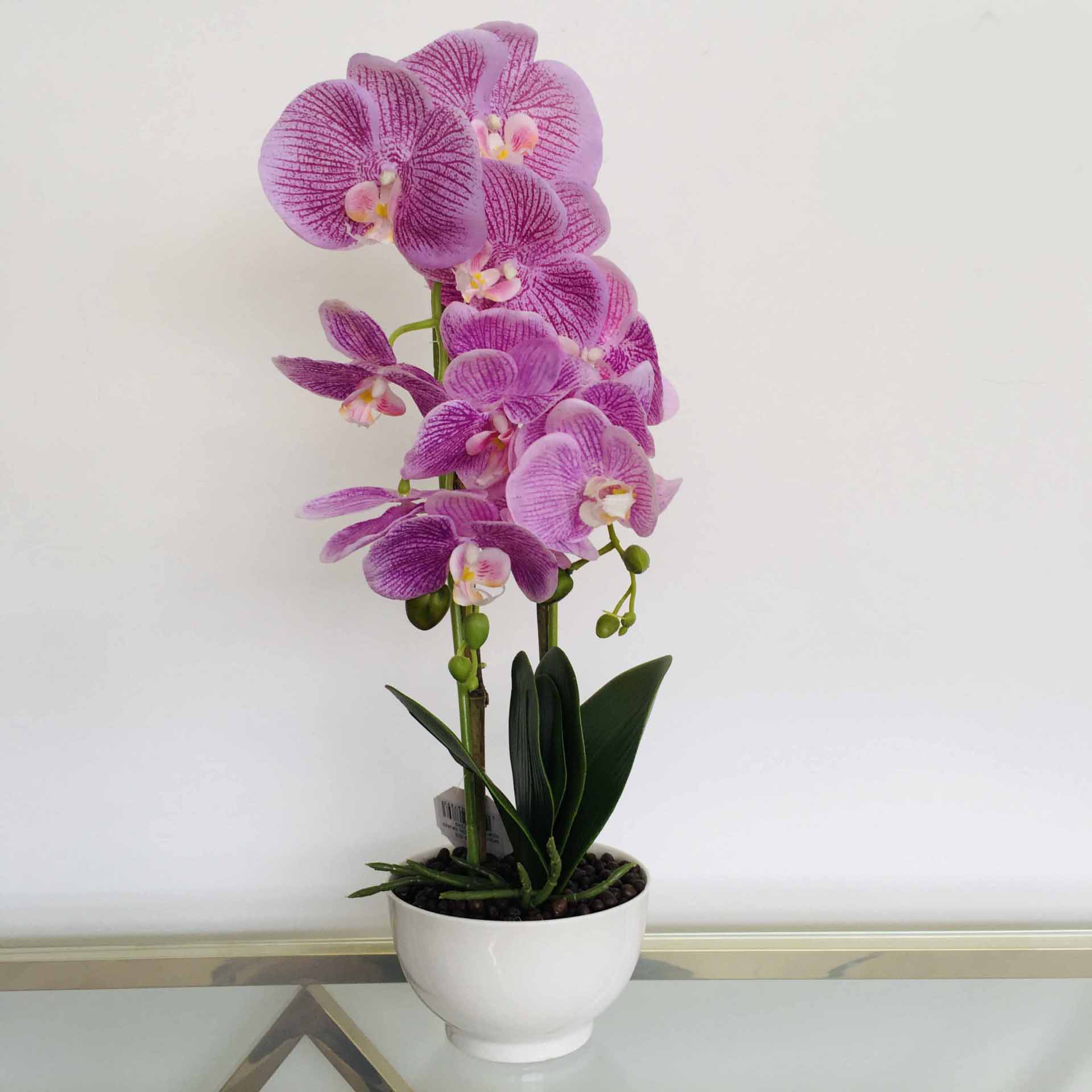 Orquídea artificial rosa em vaso branco sobre superfície espelhada.