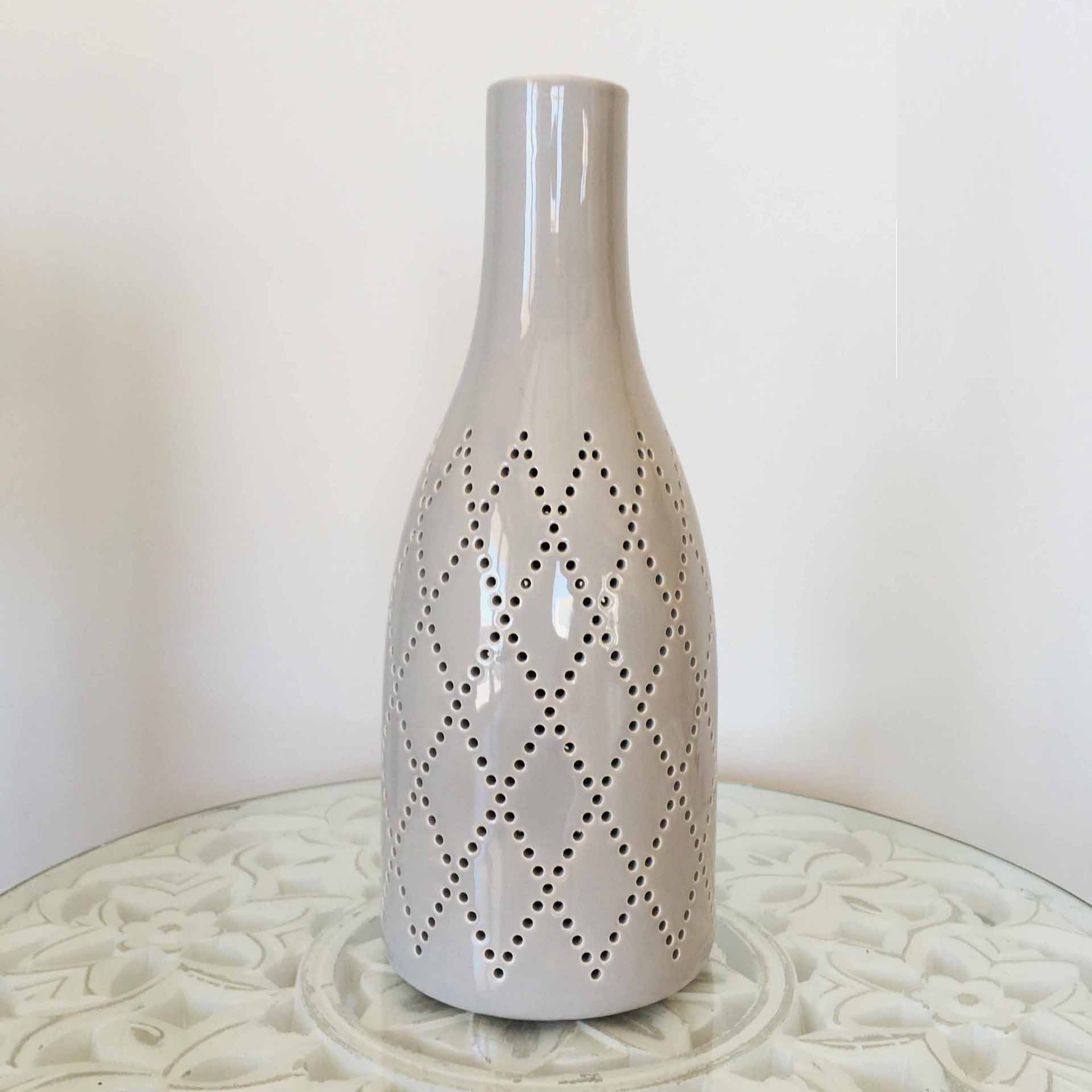 Vaso cinza claro com padrão de losangos perfurados sobre mesa branca com padrão floral.