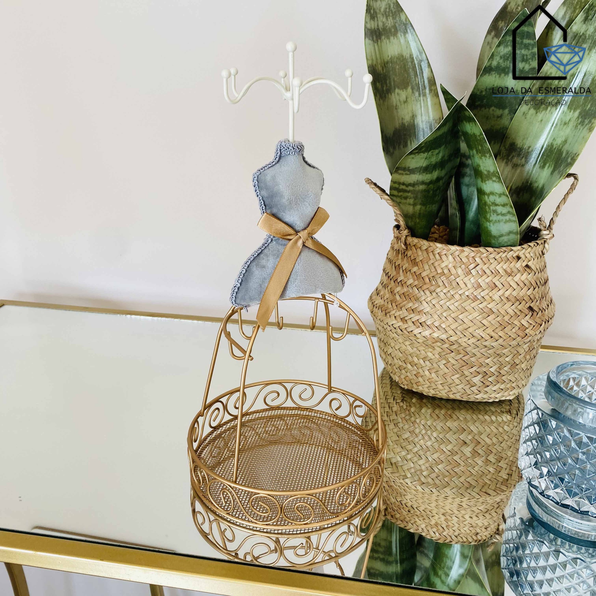 Suporte dourado para bijuterias com manequim decorativo, vaso de palhinha com planta e copo azul sobre espelho.