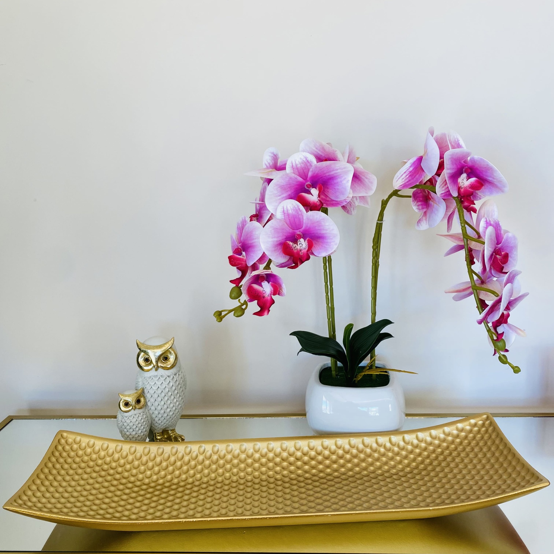 Bandeja dourada texturizada, vaso branco com orquídeas rosas e estatuetas de corujas brancas com dourado