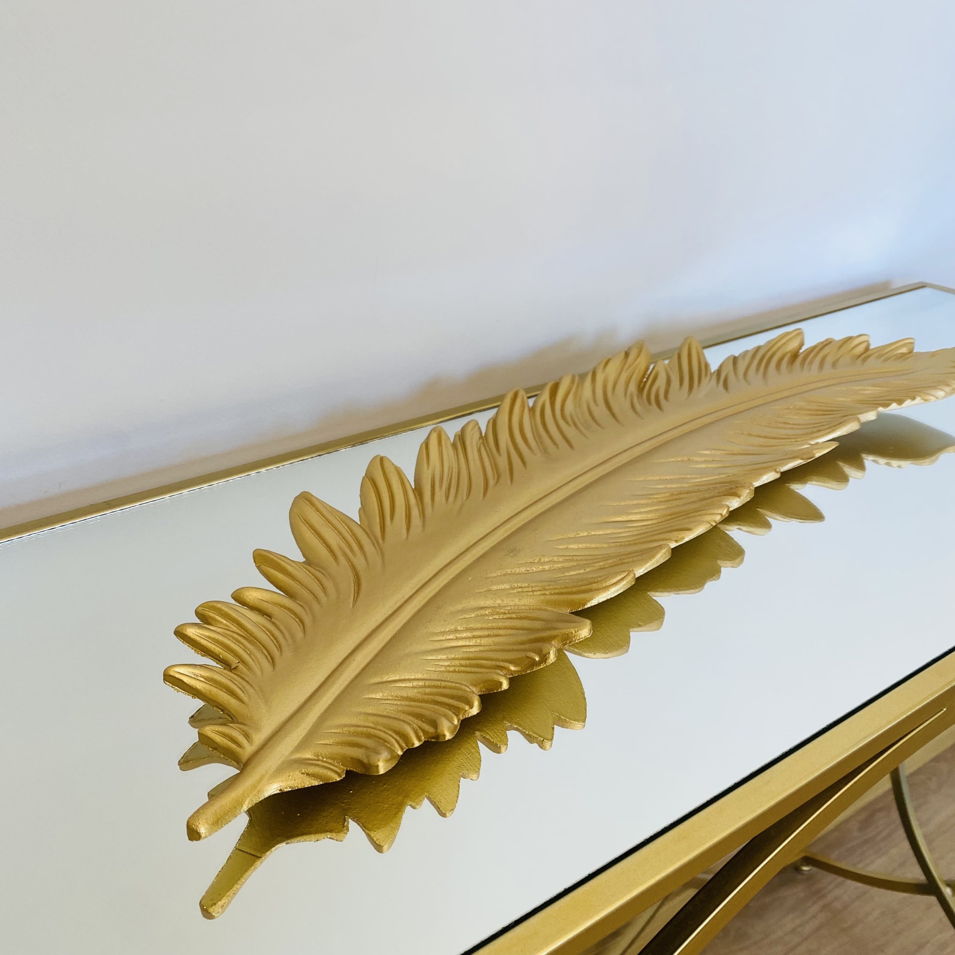 Prato decorativo dourado em forma de pena sobre mesa espelhada.