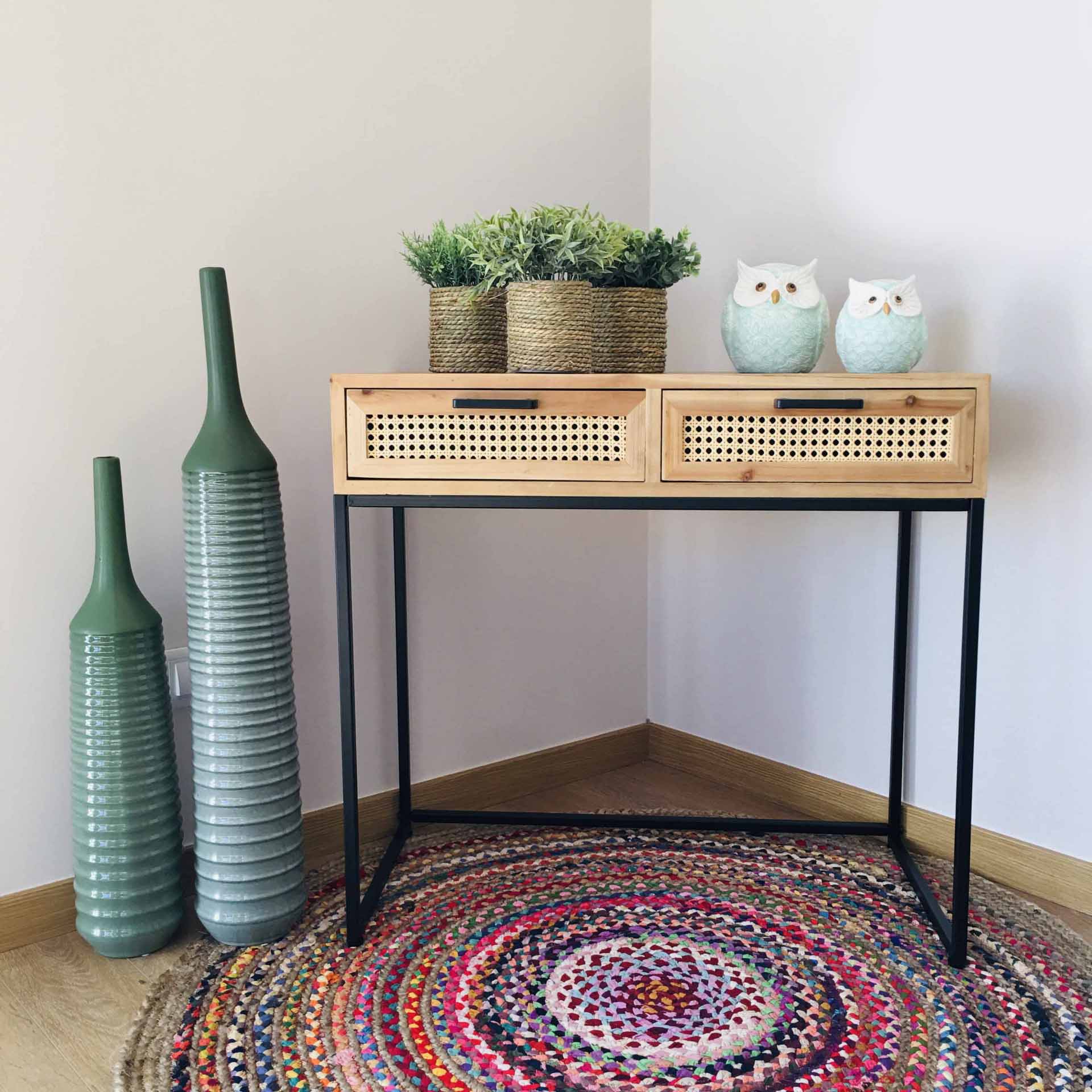Mesa de madeira clara com gavetas de palhinha, vasos verdes, corujas decorativas e tapete colorido num canto com parede branca