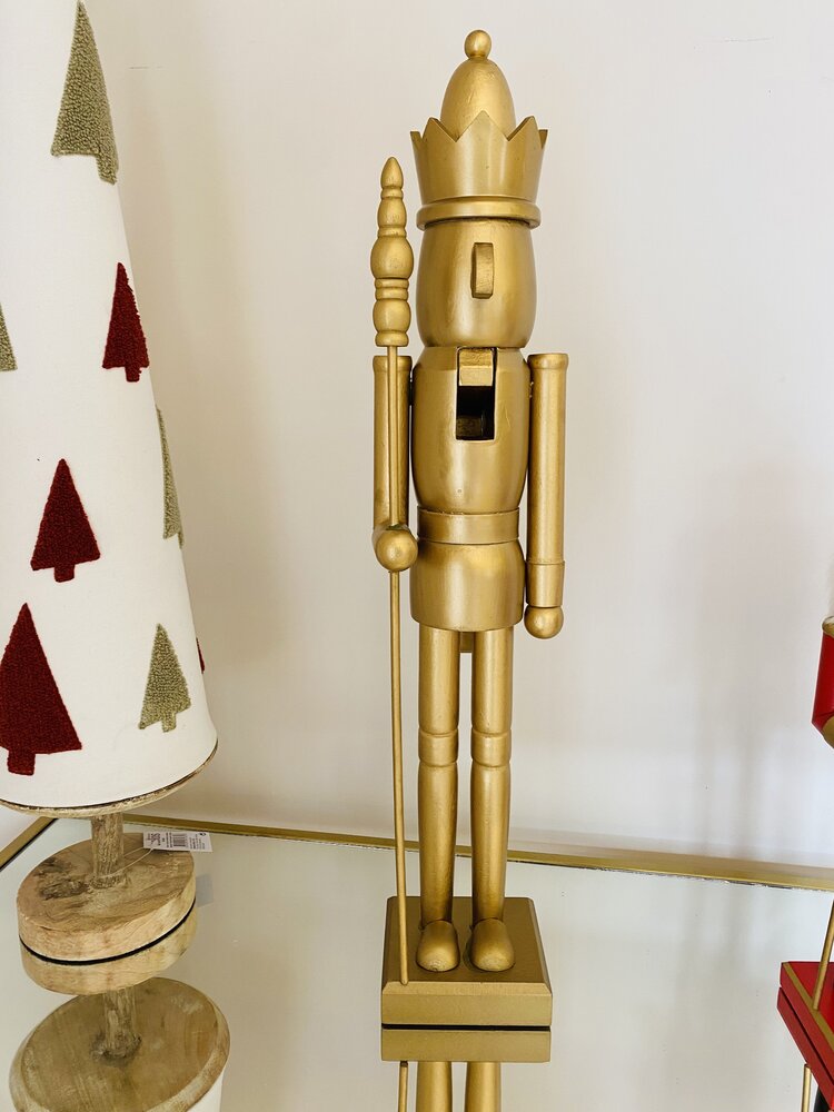 Figura dourada de soldado do rei em madeira, com coroa e lança, sobre superfície refletora ao lado de árvores de Natal decorativas.