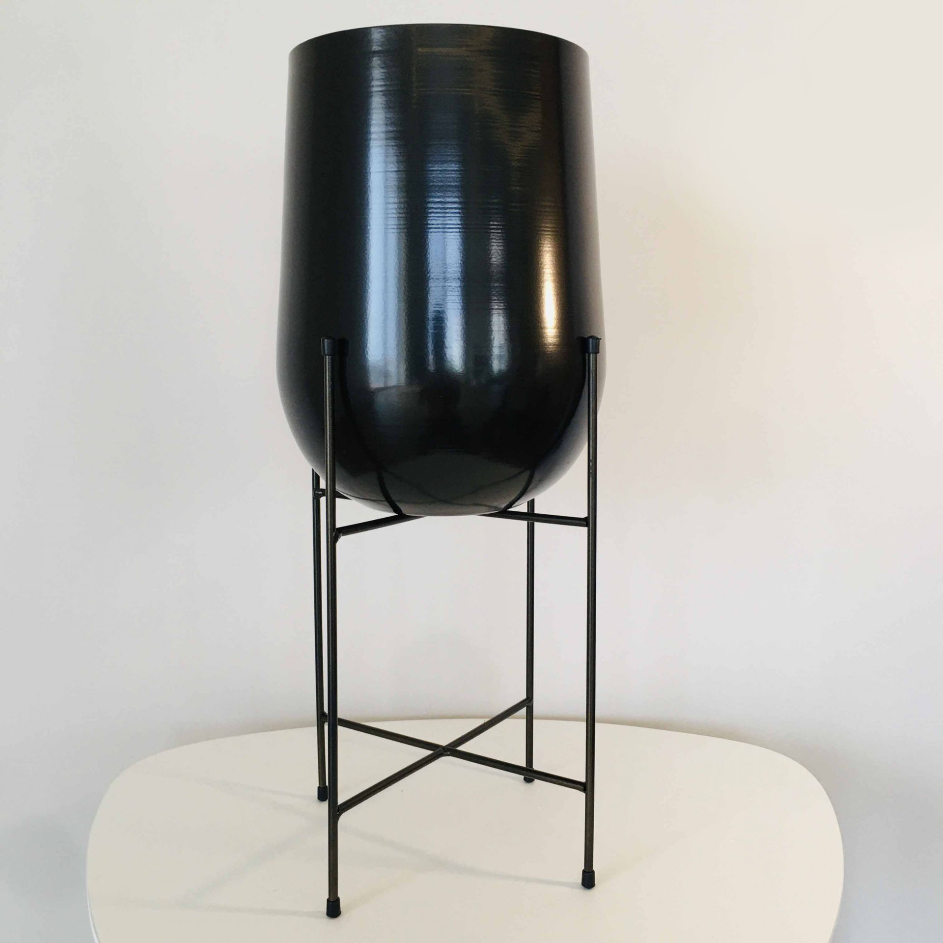 Vaso de planta alto em metal preto com suporte de pernas finas sobre mesa branca