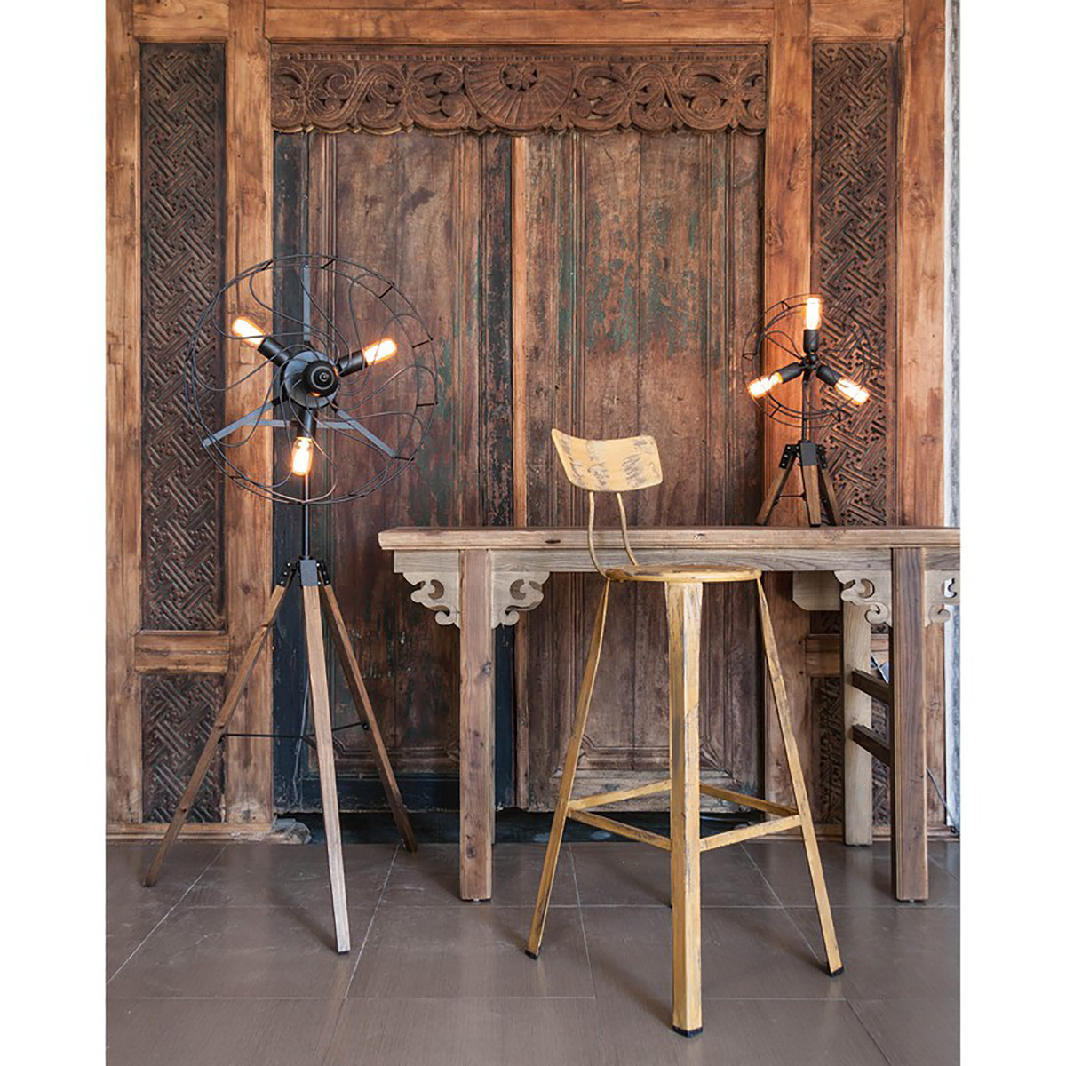 Lâmpadas vintage em tripés de madeira ao lado de mesa e banco altos em madeira e metal, frente a portas de madeira trabalhada.