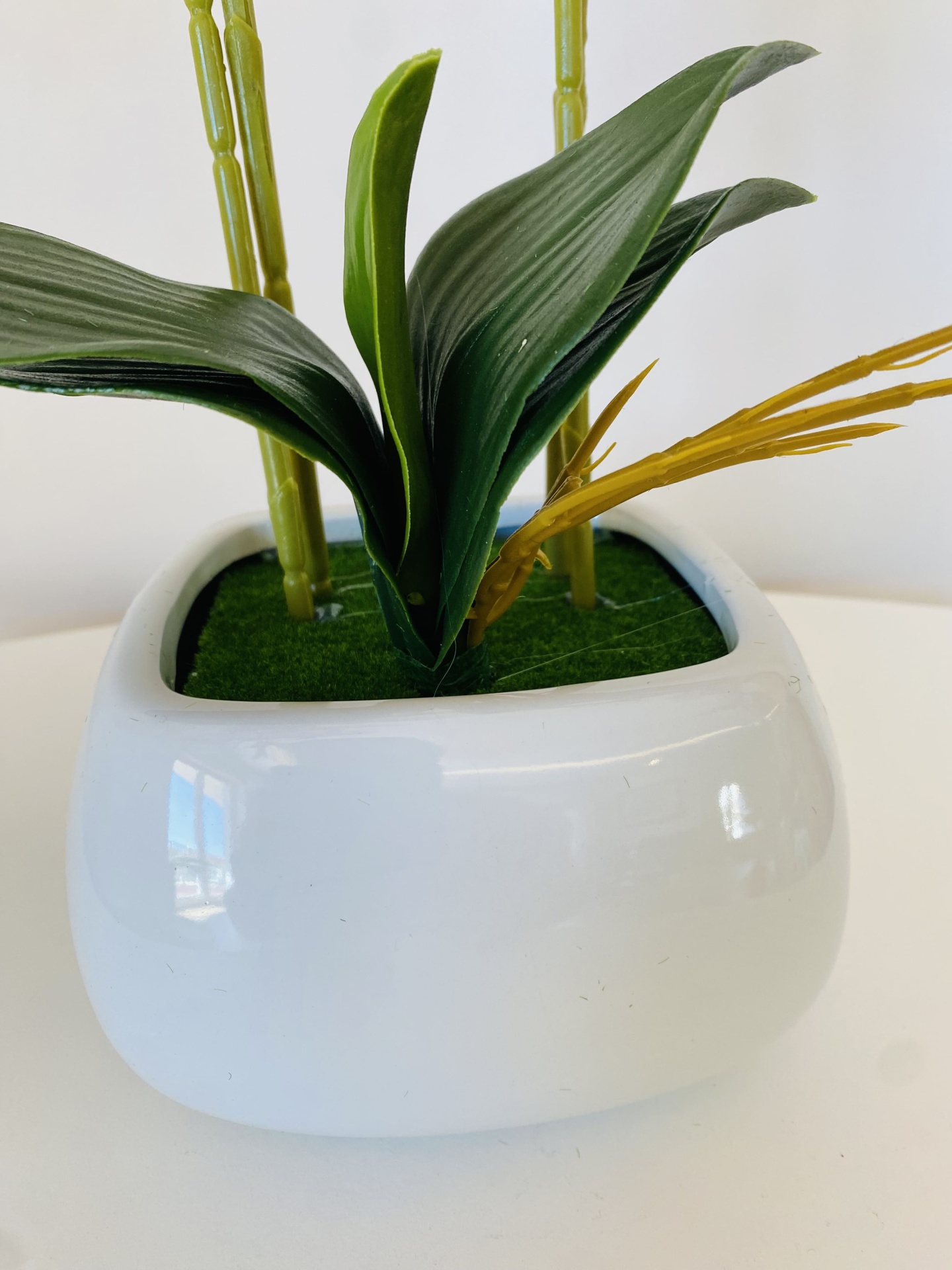 Vaso branco com planta artificial de folhas verdes e caules amarelos.