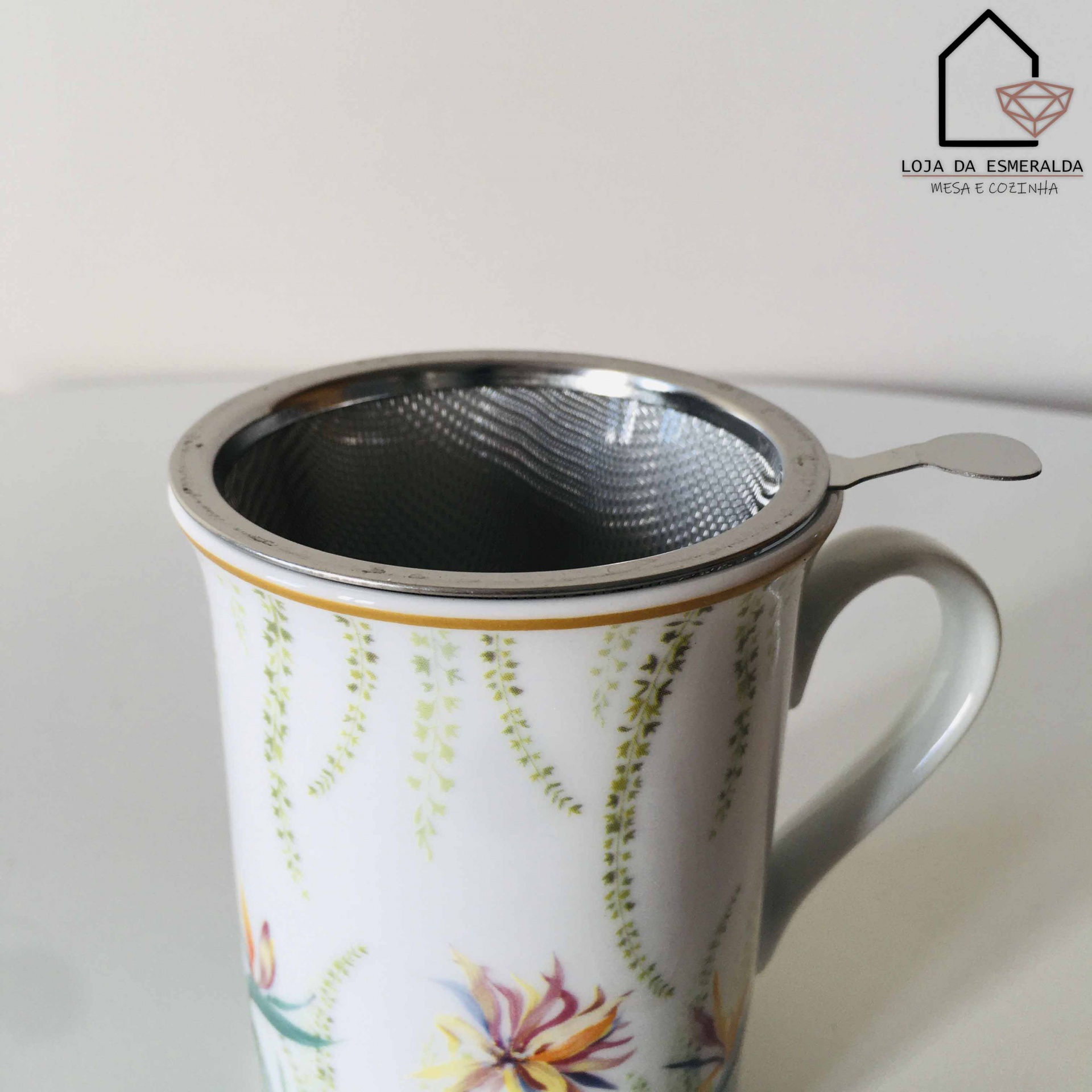 Caneca branca decorada com flores e infusor metálico em superfície branca.