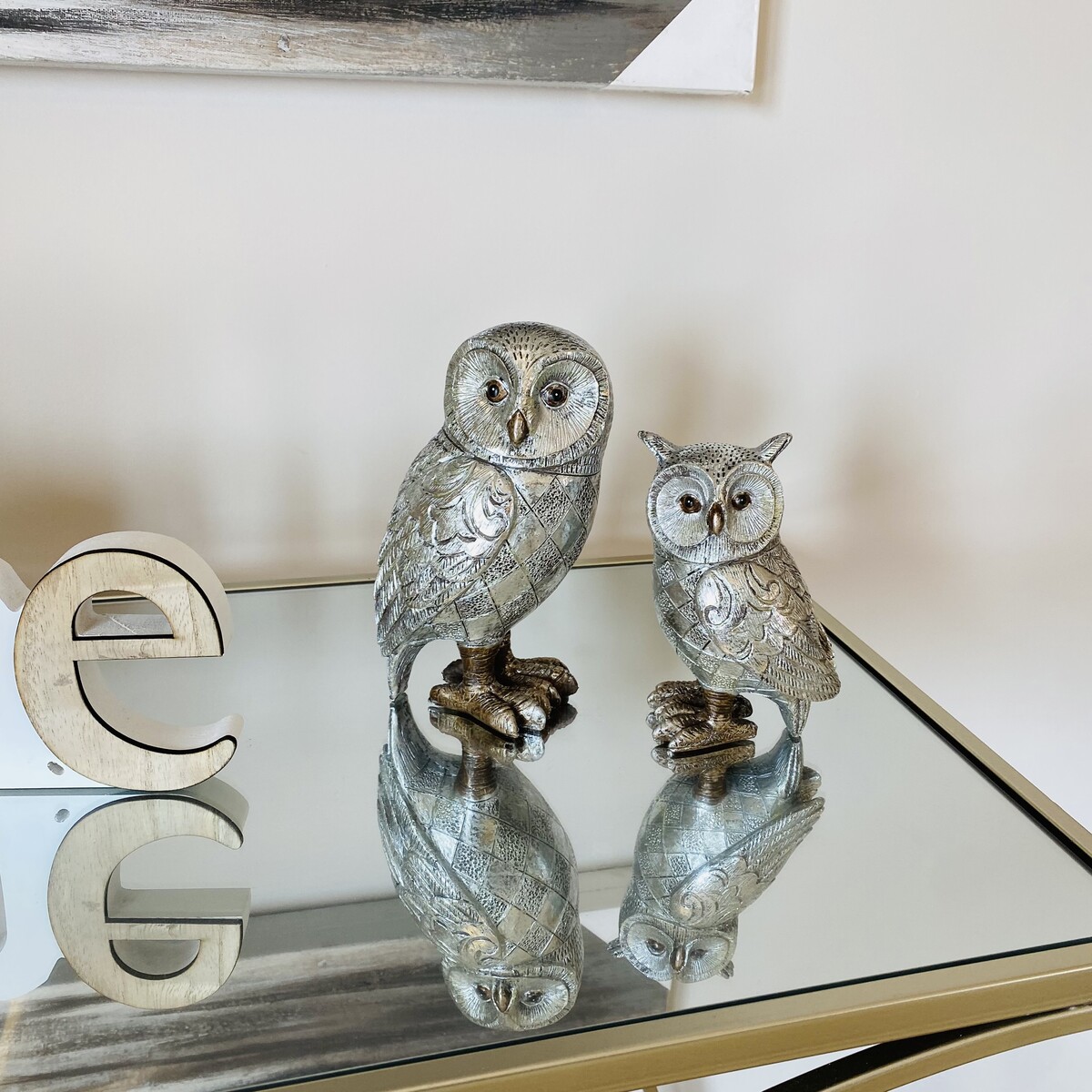 Duas esculturas de coruja prateadas sobre mesa espelhada com letra decorativa
