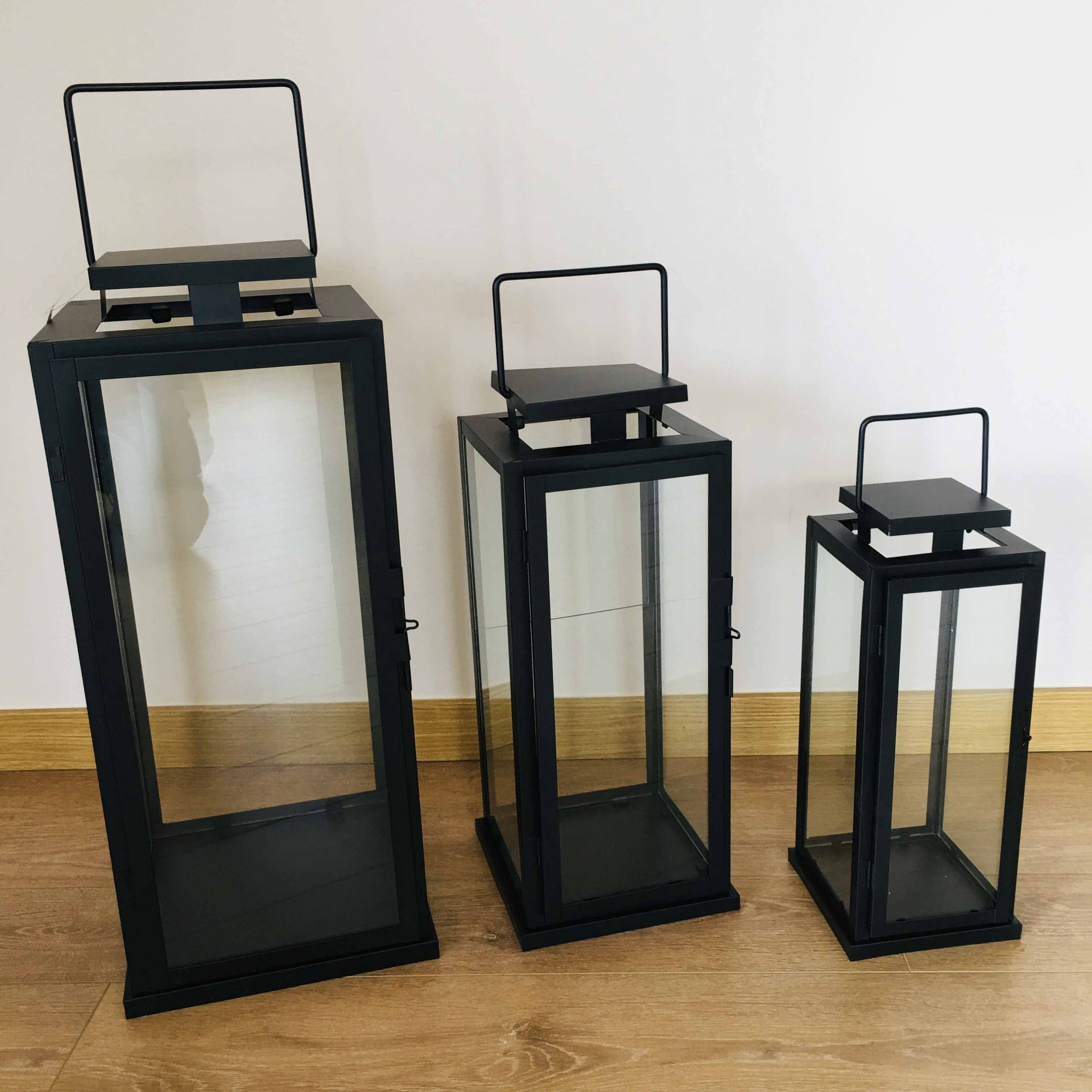 Três lanternas pretas de metal e vidro de vários tamanhos alinhadas no chão junto a uma parede branca