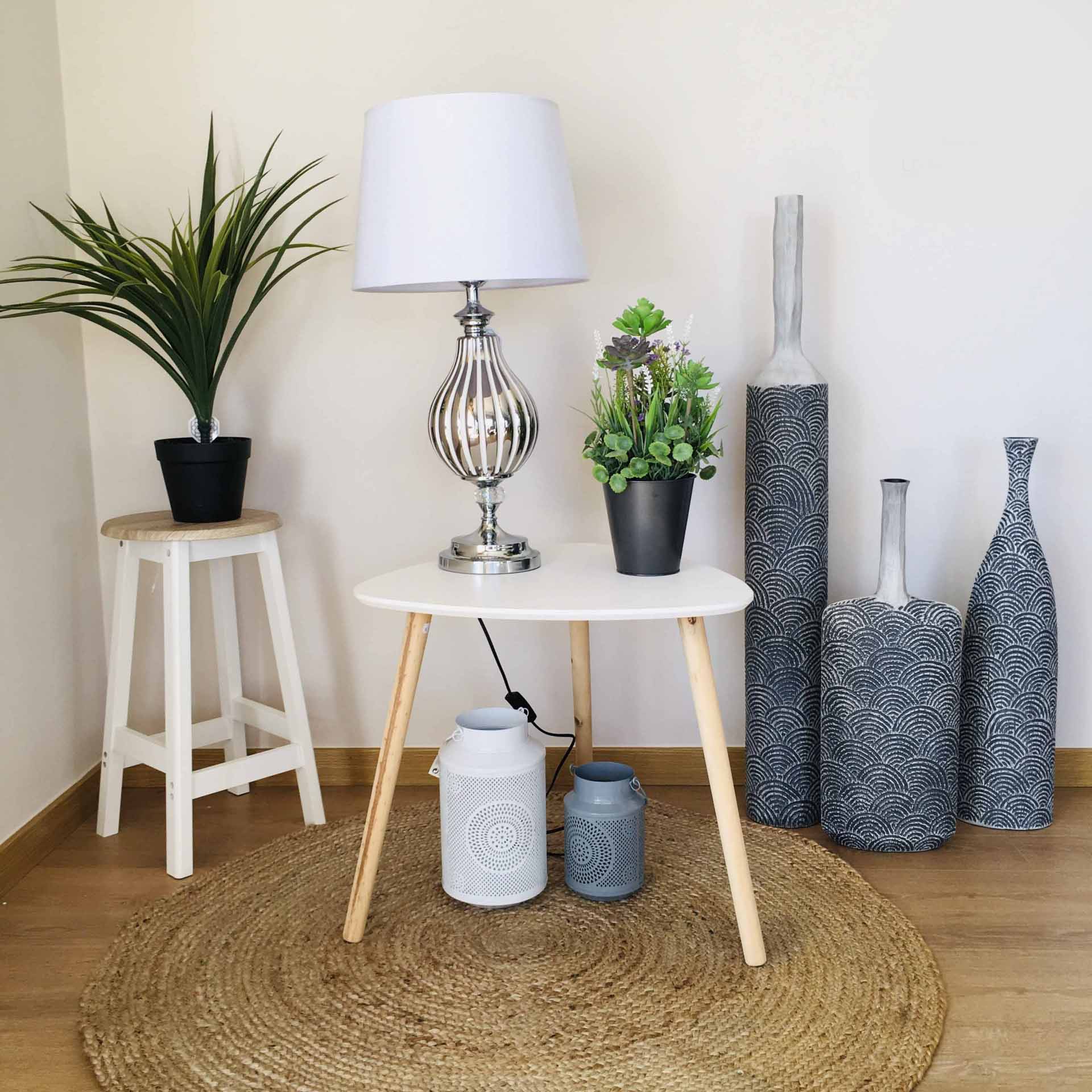 Mesa branca redonda com candeeiro e plantas decorativas num ambiente interior com chão de madeira e tapete de juta