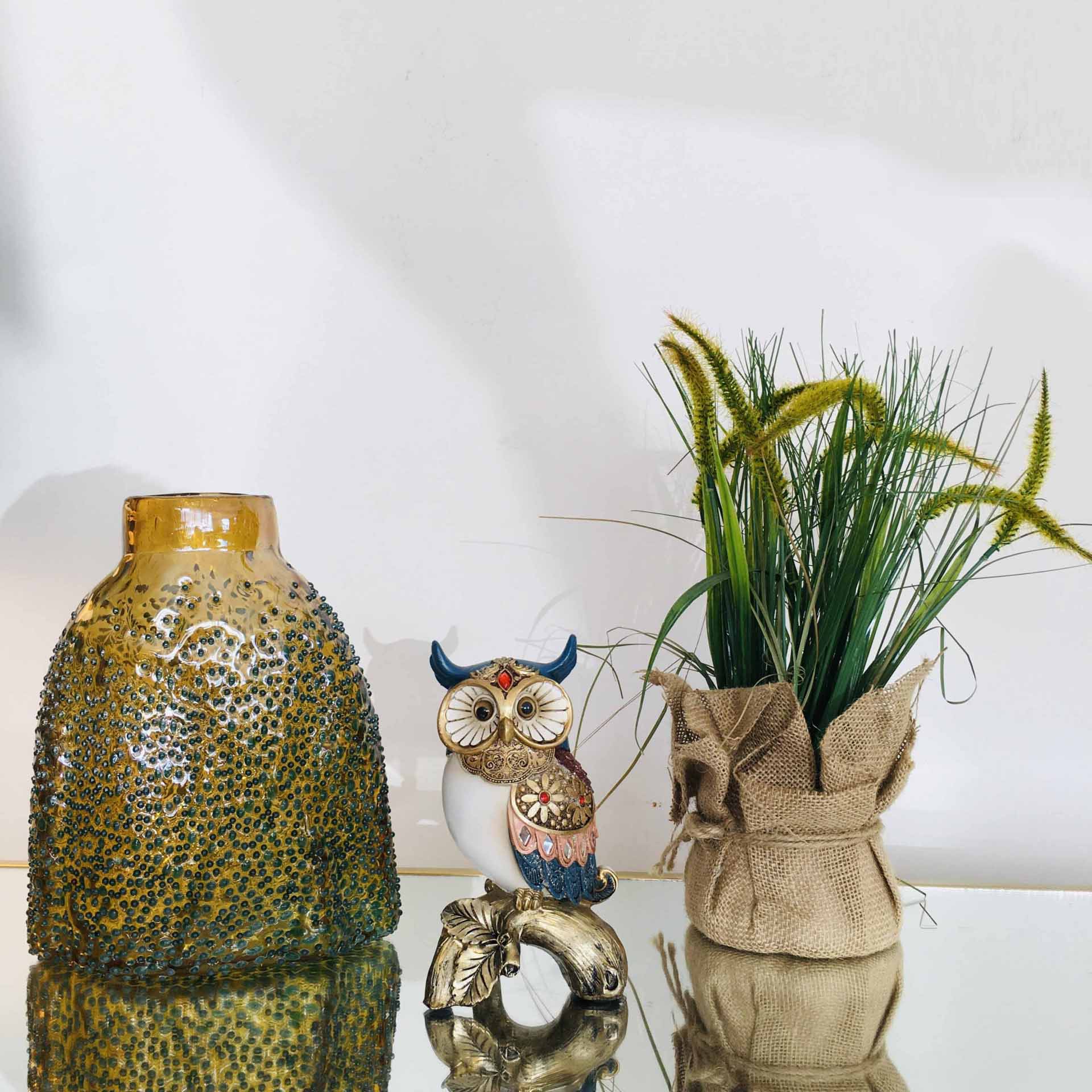 Vaso decorativo verde, coruja cerâmica e planta artificial em vaso de tecido sobre superfície refletora.