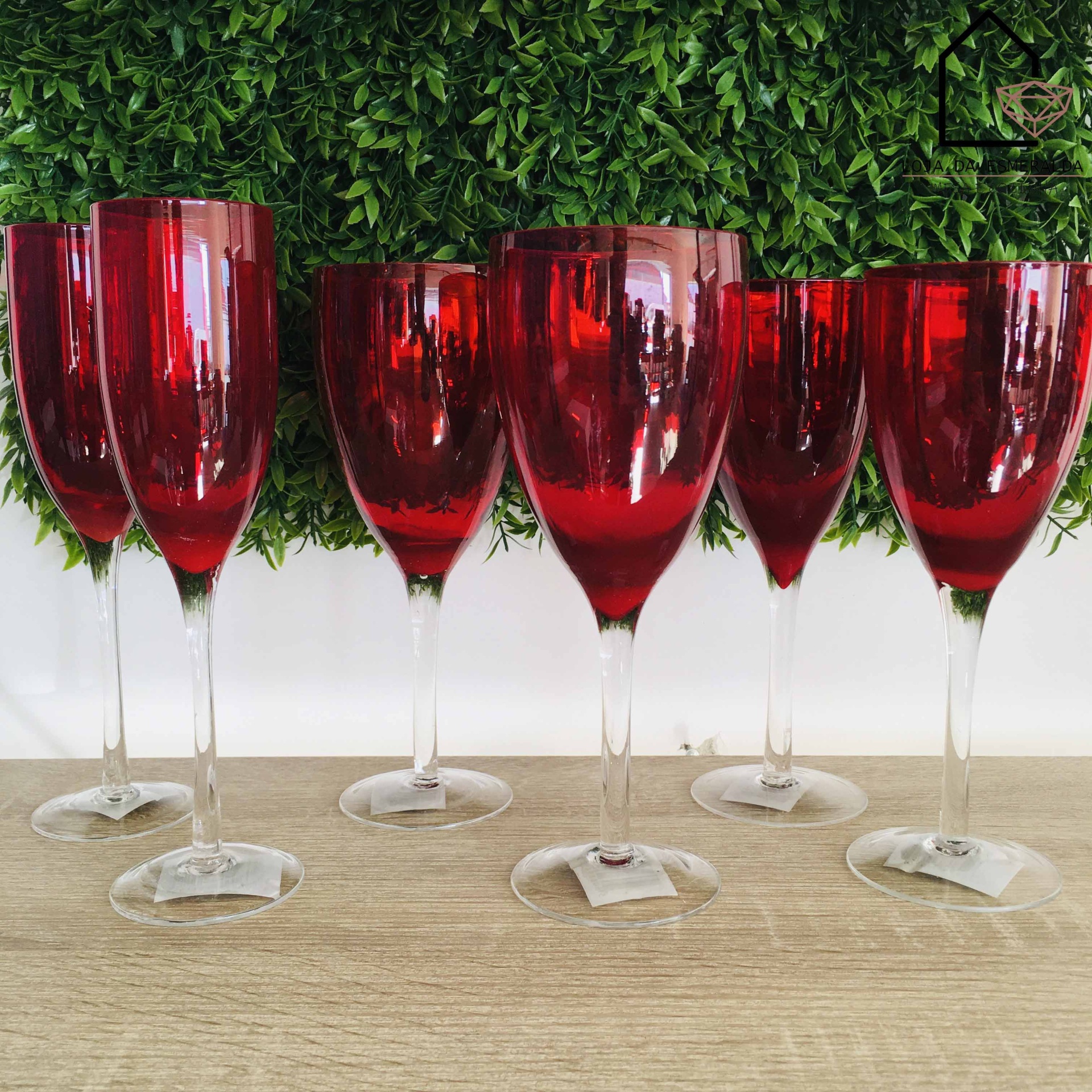 Seis copos de vinho vermelhos translúcidos com hastes claras sobre mesa de madeira com fundo de folhas verdes