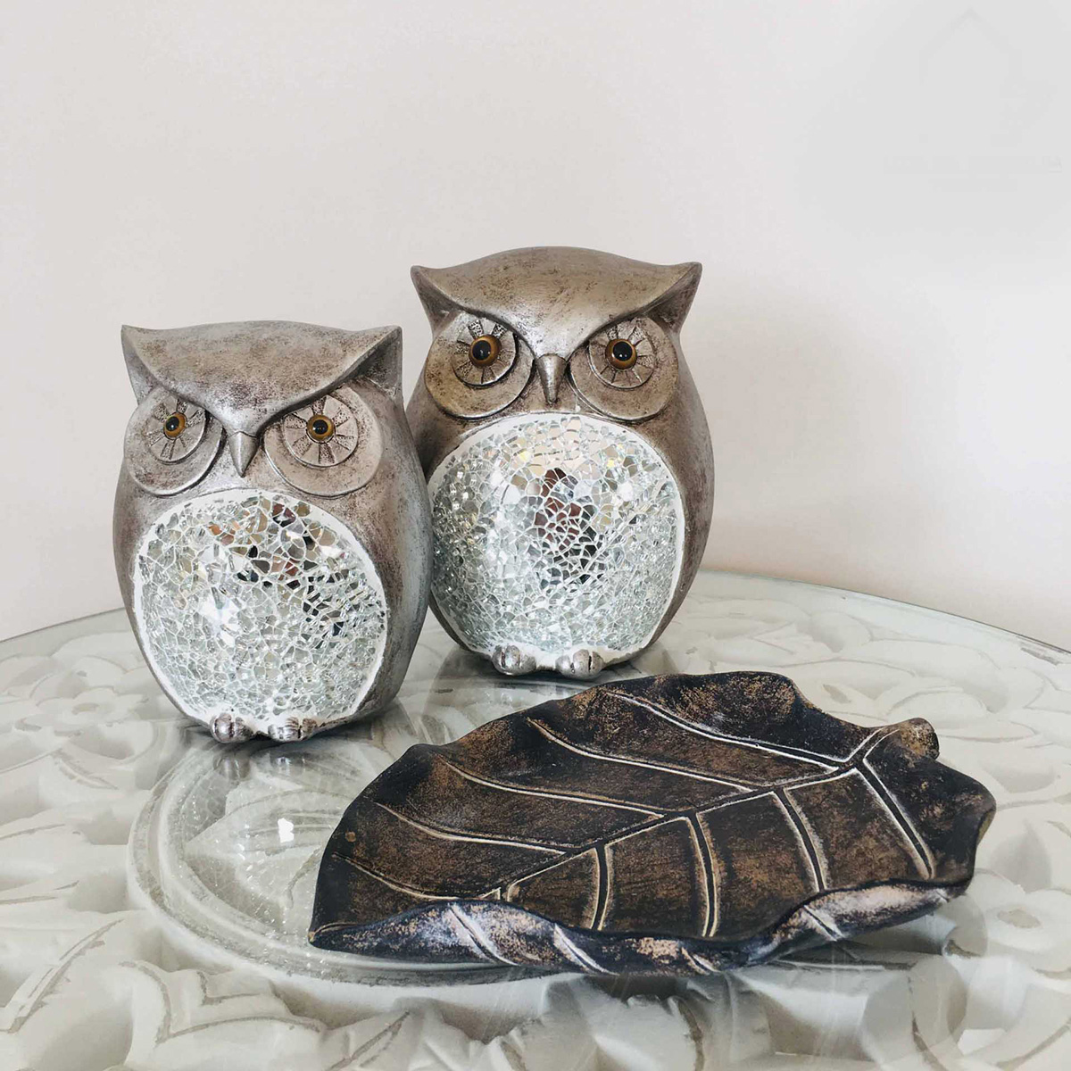 Duas corujas decorativas de cerâmica com detalhes prateados e uma peça em forma de folha escura sobre uma superfície de vidro