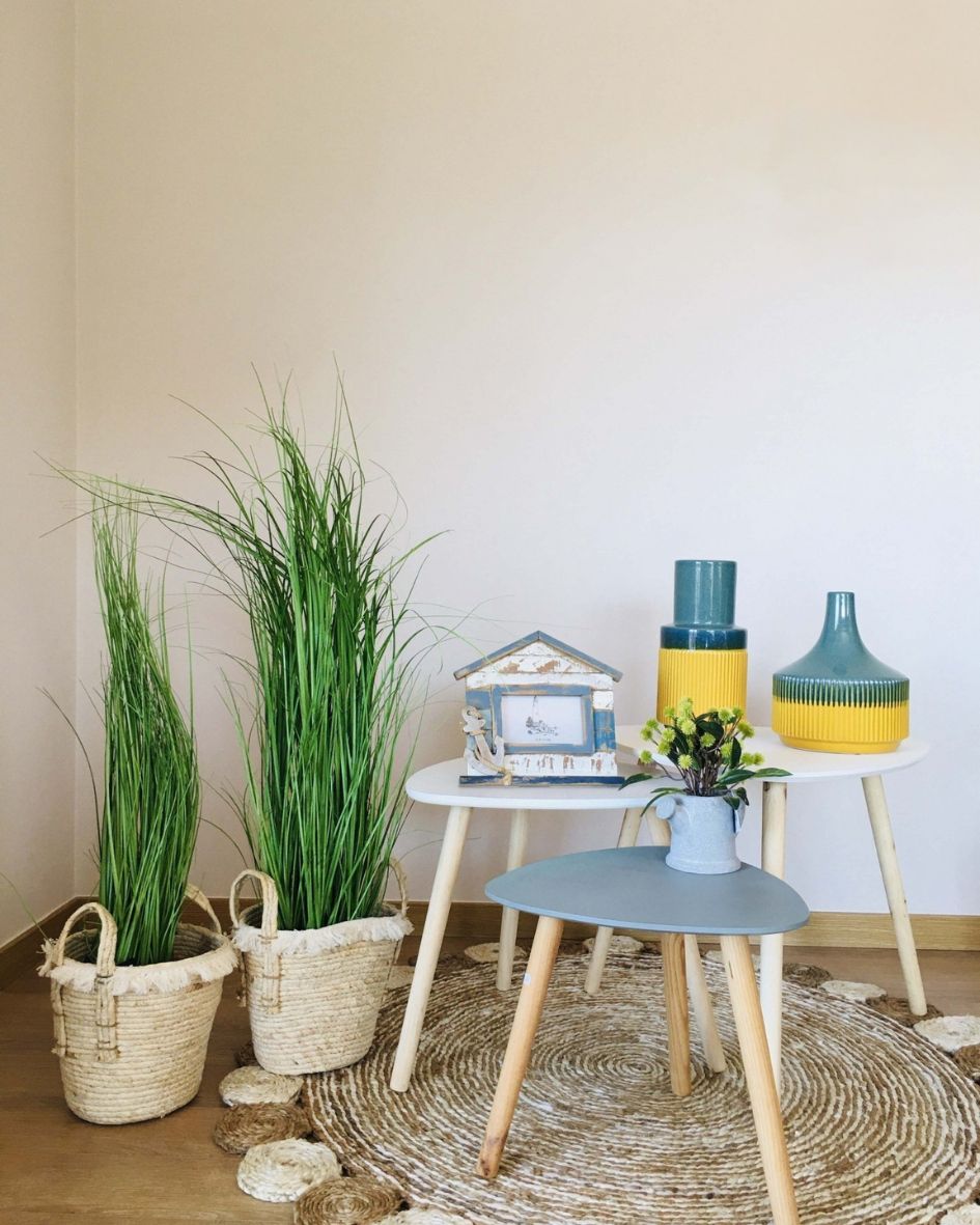 Vasos de palha com plantas verdes, mesas pequenas de madeira com objetos decorativos azul e amarelo em tapete redondo