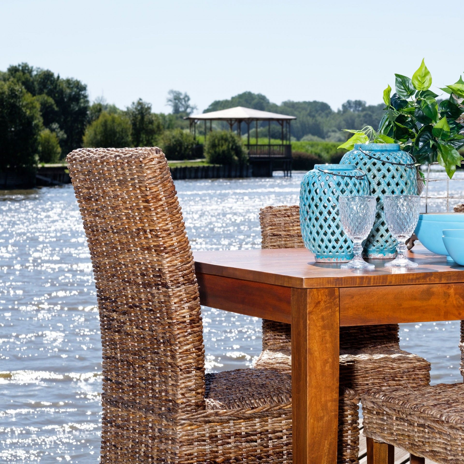 Mesa de madeira e cadeiras de fibras naturais junto a um lago com vasos azuis e copos de vidro.