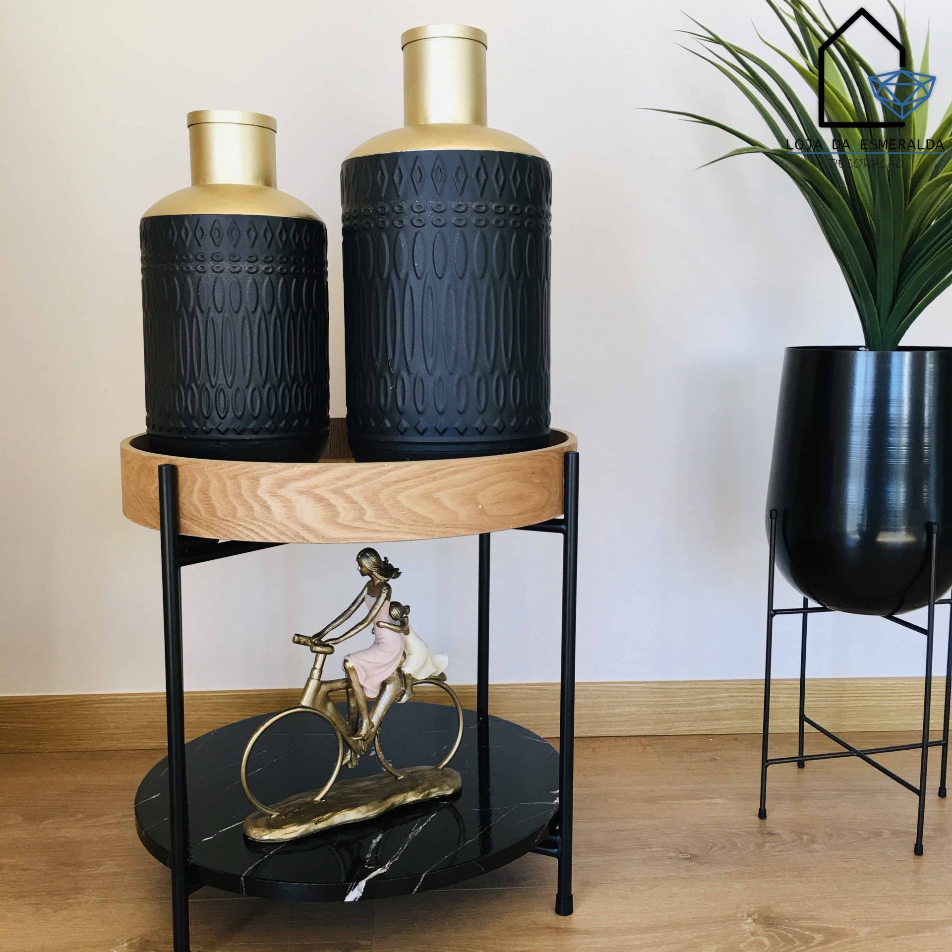 dois jarros decorativos preto e dourado em suporte redondo ao lado de vaso preto com planta
