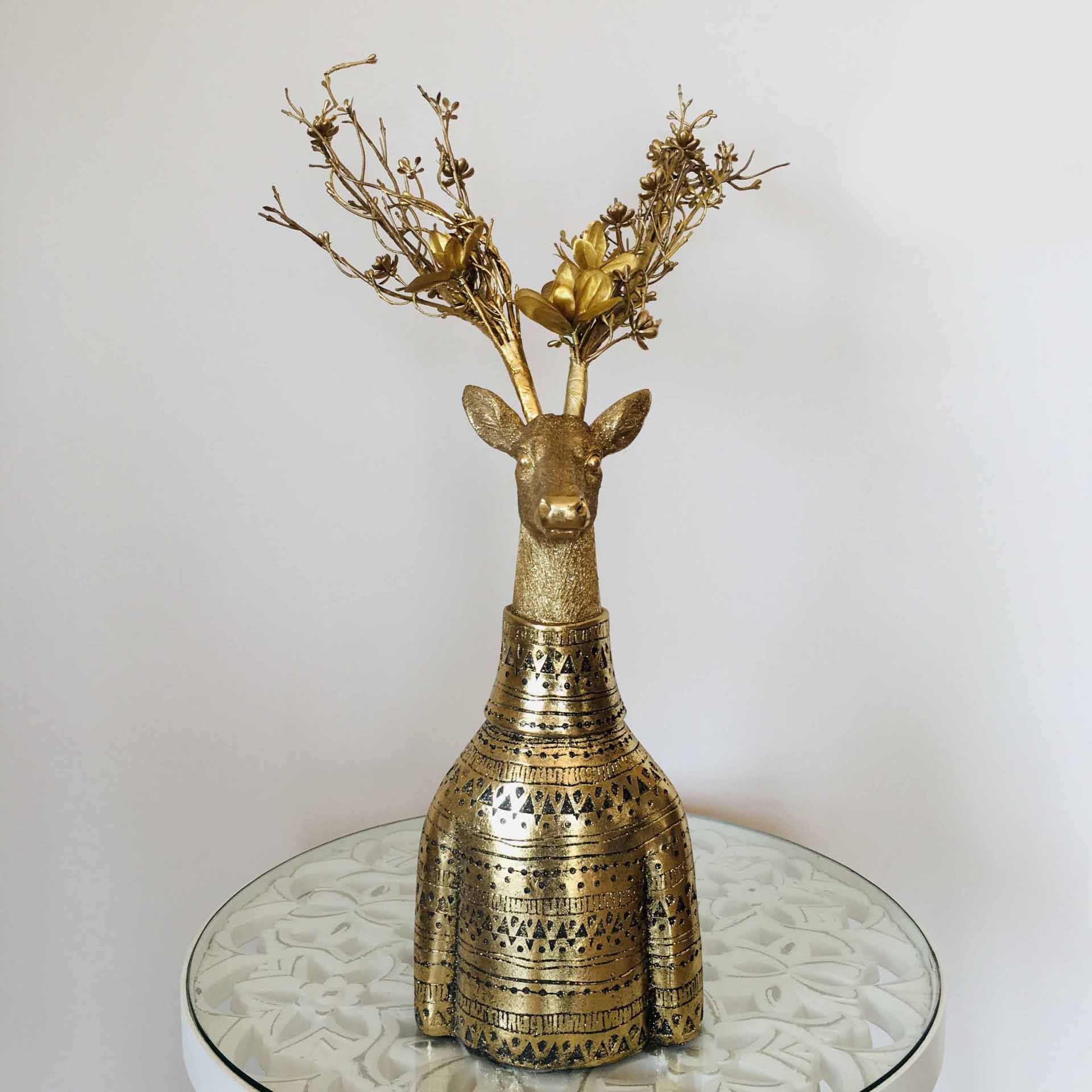 Escultura dourada de veado com galhos secos e flores colocada num tampo espelhado